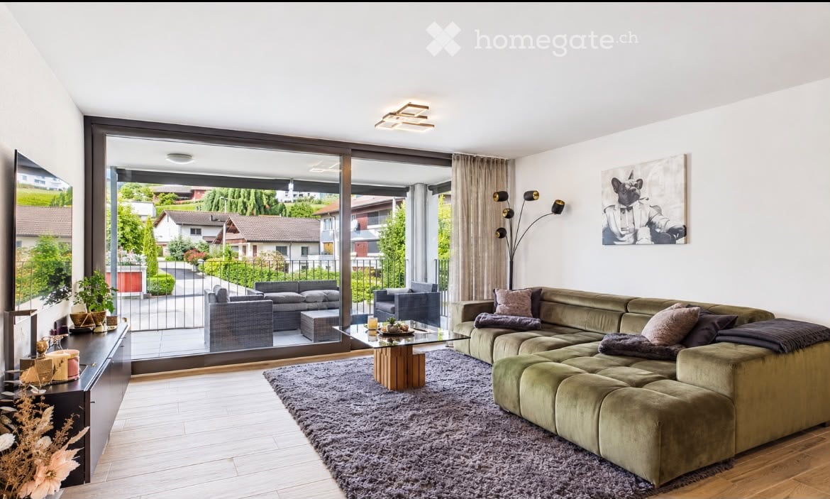 Modern living room with large windows offering a view, featuring a sectional sofa, coffee table, side table, floor lamp, and decorative plants.