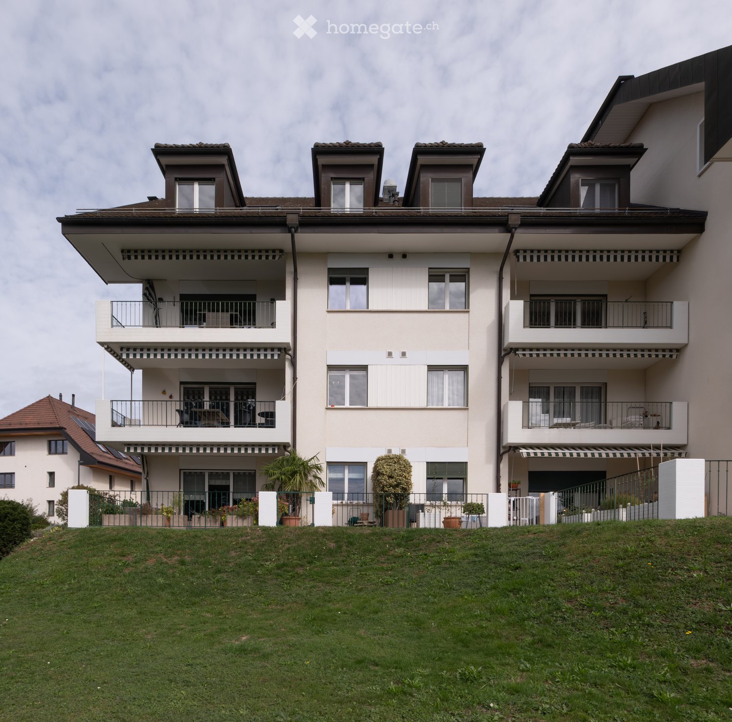 A multi-story residential building with balconies, large windows, and a raised ground floor. The building features a modern design with beige and brown tones, surrounded by a well-maintained lawn and potted plants.