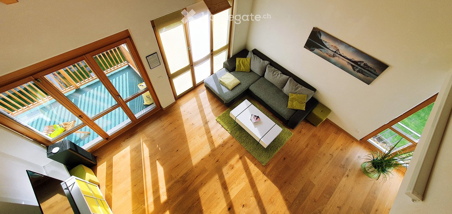 Modern living room with wooden flooring, large windows, and a balcony. Features a grey sectional sofa with yellow cushions, a white coffee table, and a green ottoman.