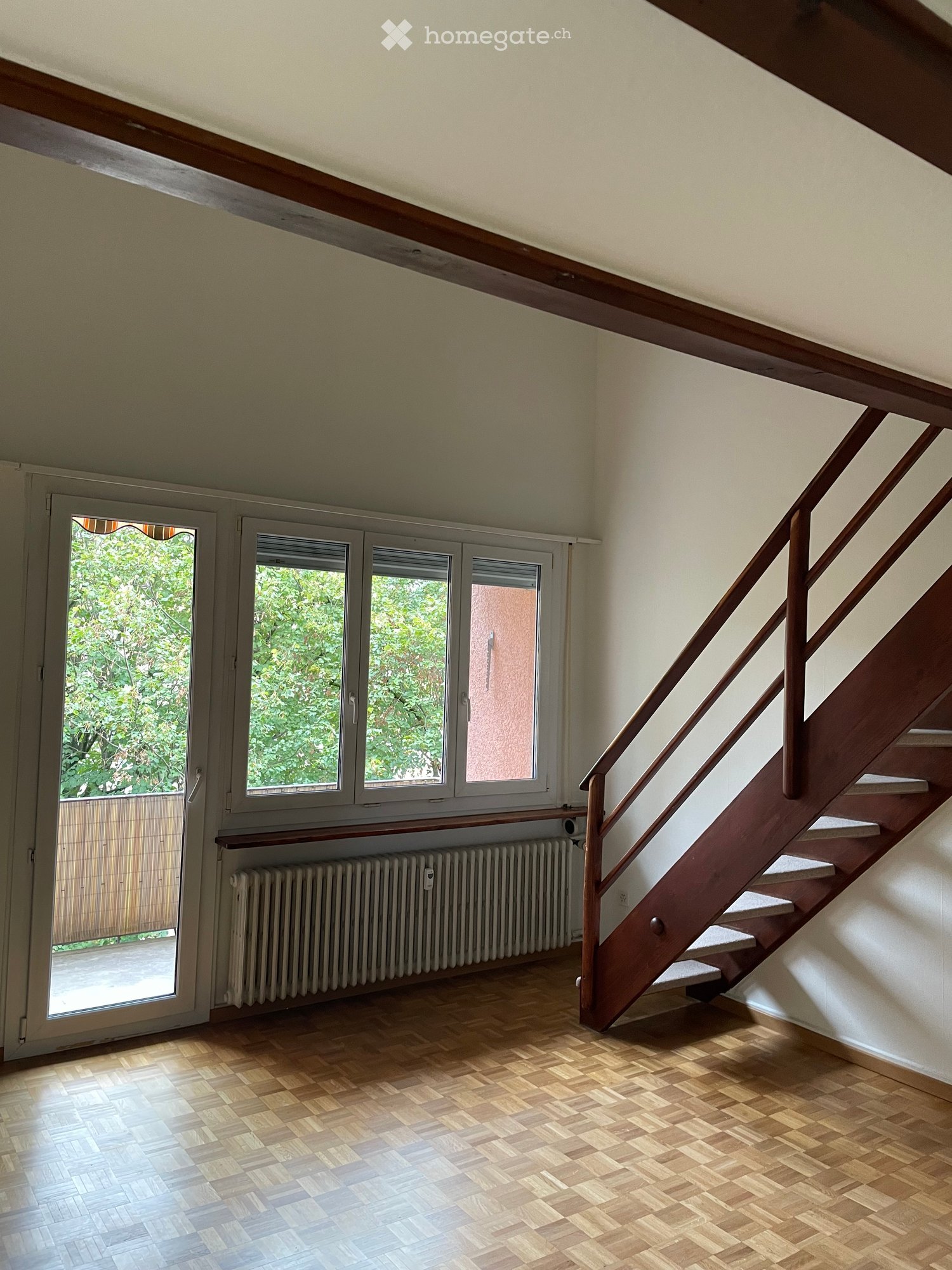 Empty hallway with wooden flooring, white walls, a staircase with wooden railings, and large windows offering a view of greenery outside.