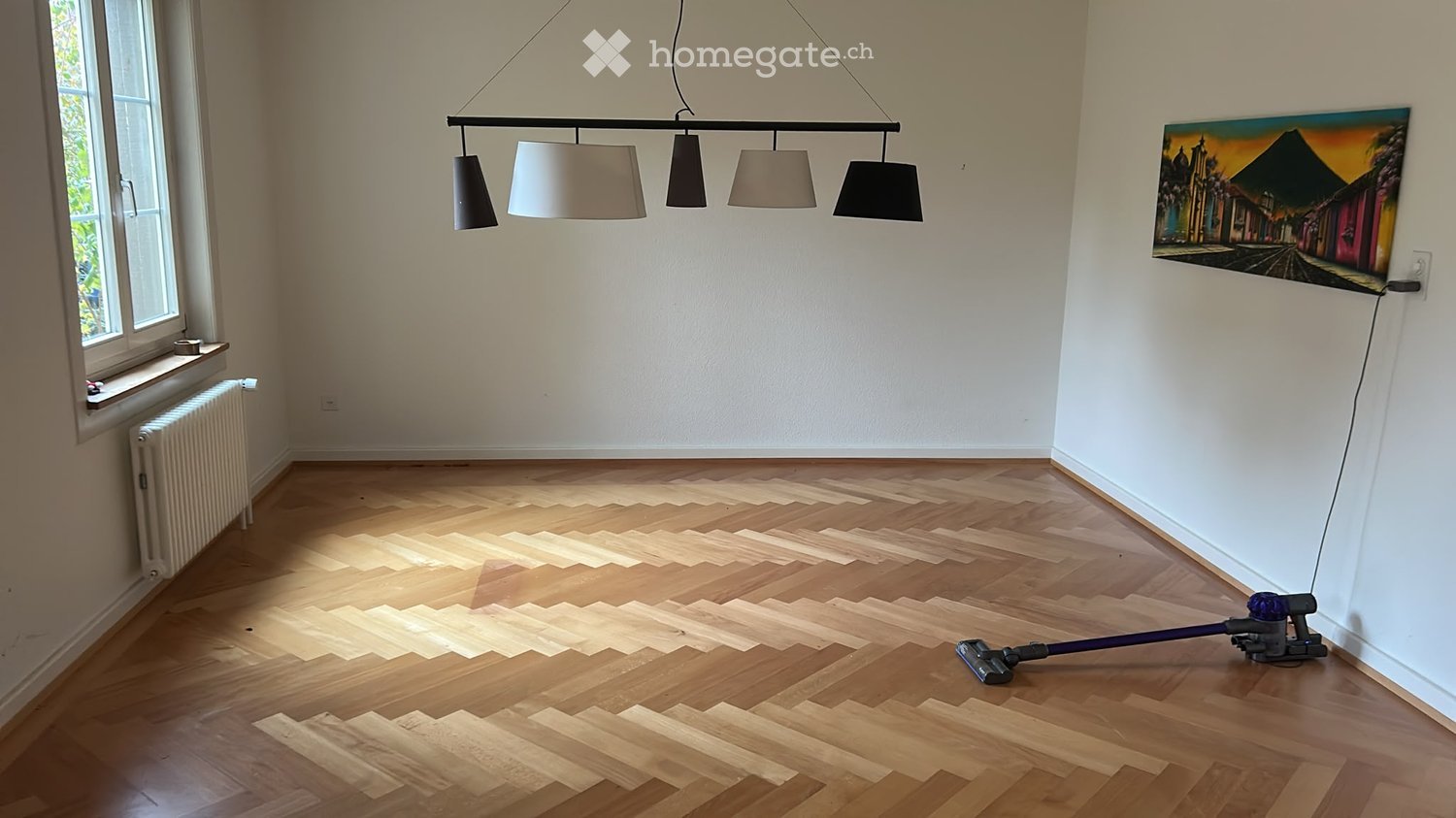 Empty room with wooden flooring, white walls, large window, radiator, and a painting on the wall. A vacuum cleaner is placed on the floor.