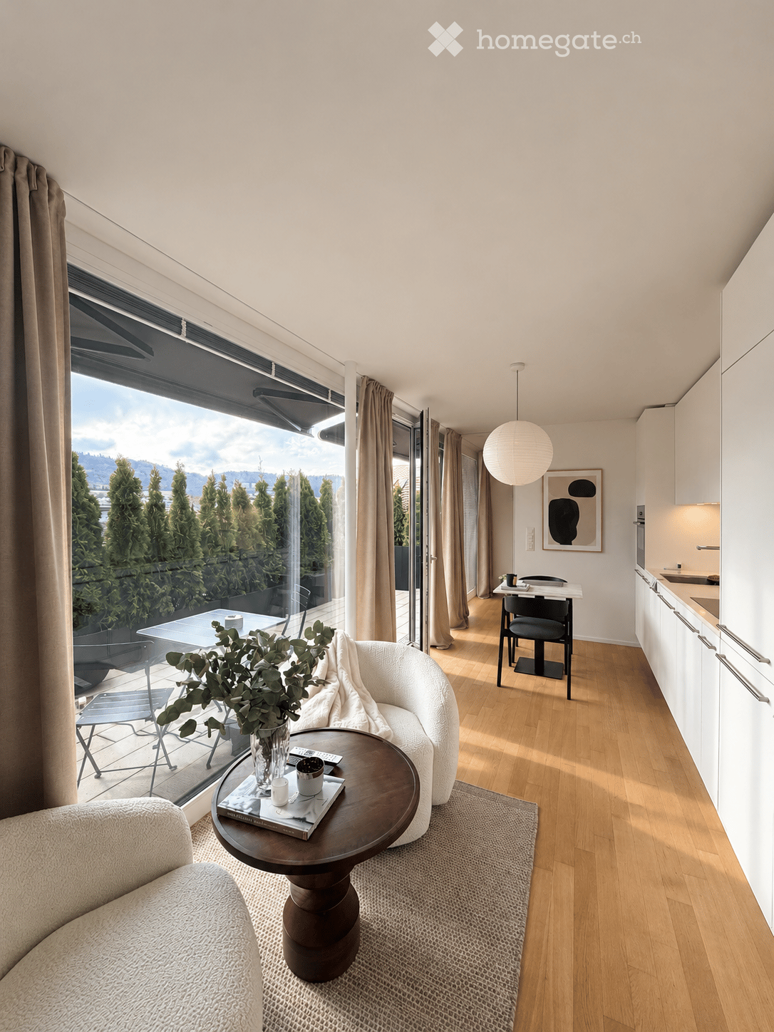 Modern living room with large windows offering a scenic view, featuring a white sofa, round coffee table, dining area with chairs, and a small kitchen area with cabinets.
