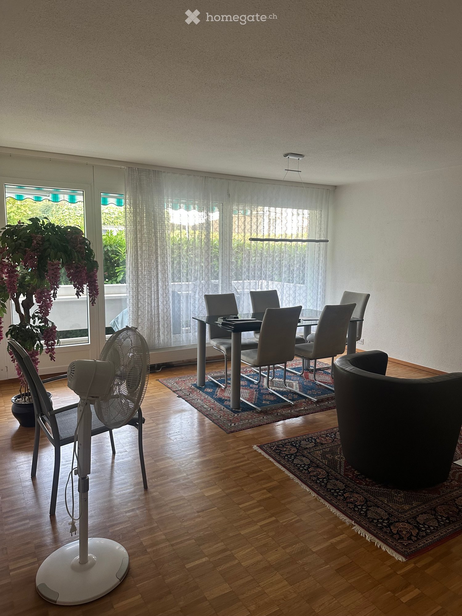 A modern living room with wooden flooring, a large window with sheer curtains, a dining table with six chairs, a black leather sofa, a standing fan, and a potted plant near the window.
