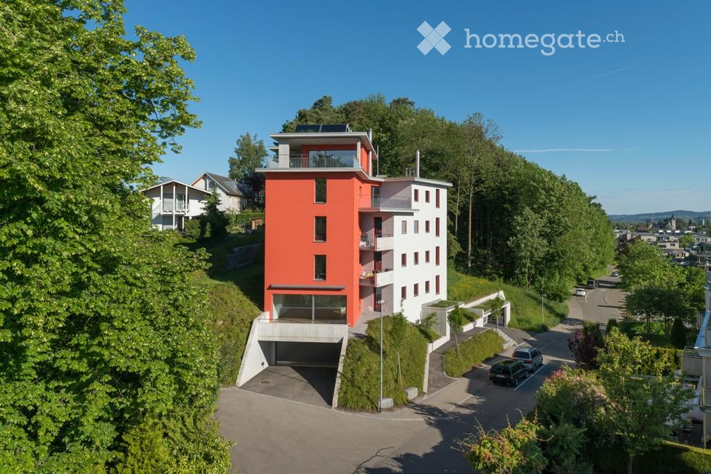 Modern multi-story building with red and white facade, balconies, and integrated garage. Surrounded by greenery and parking area.