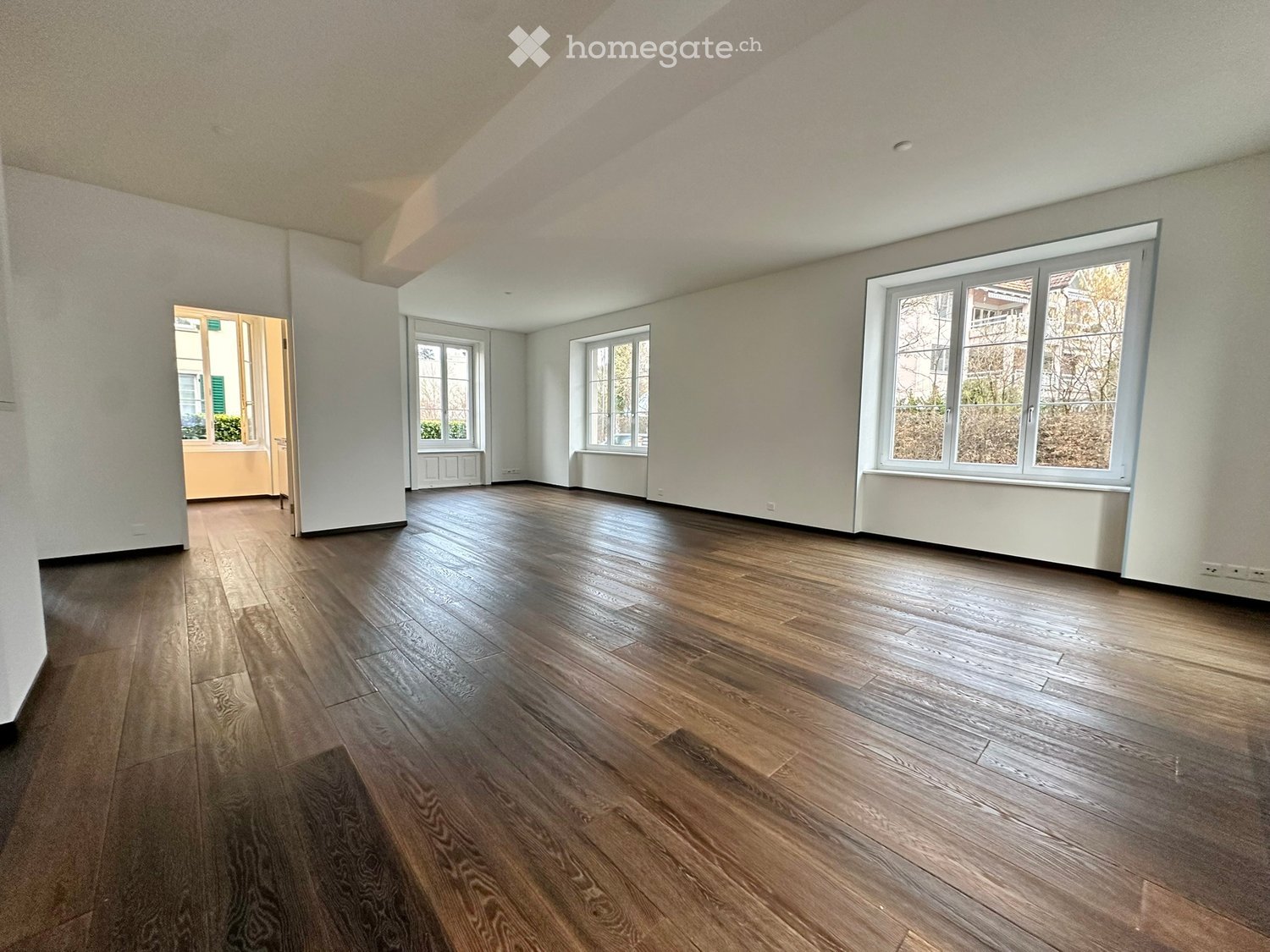 Empty room with wooden flooring, white walls, and multiple windows allowing natural light. The space is completely unfurnished with no visible furniture or equipment.