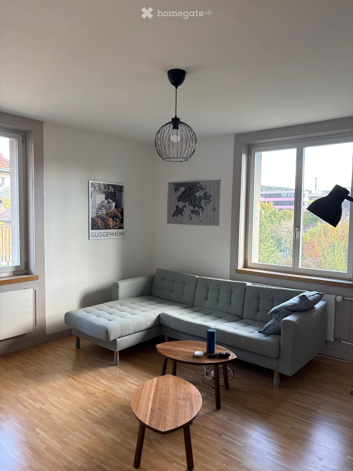 Modern living room with light grey sectional sofa, wooden coffee table, small round table, pendant lamp, and artwork on the walls.