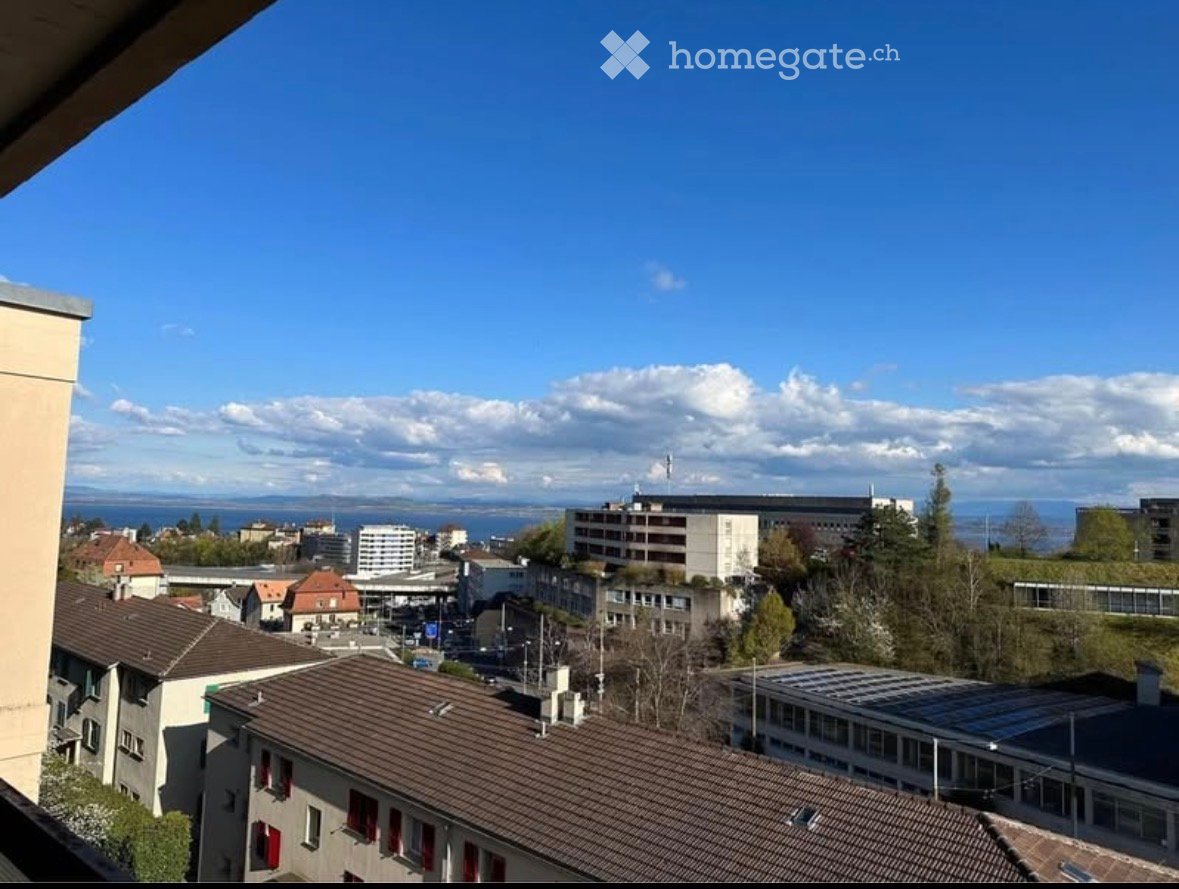 Panoramic cityscape view from a balcony, featuring rooftops, buildings, and distant water body under a clear blue sky.
