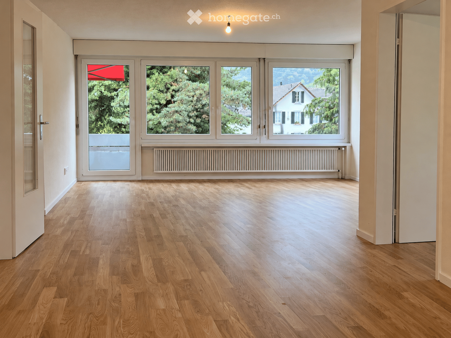 Empty room with wooden flooring, large windows, radiator, and a door leading to an outdoor area.