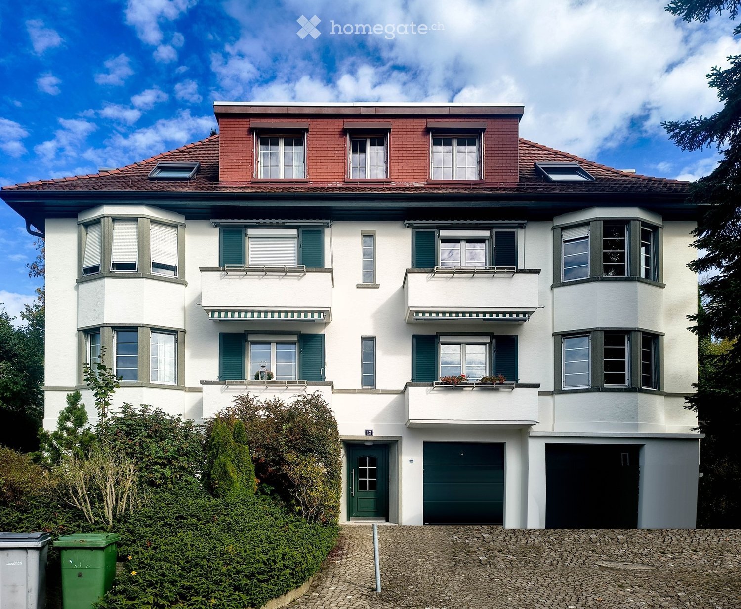 A two-story house with white walls, green shutters, and a red-tiled roof. Features include multiple windows, balconies, and a garage.