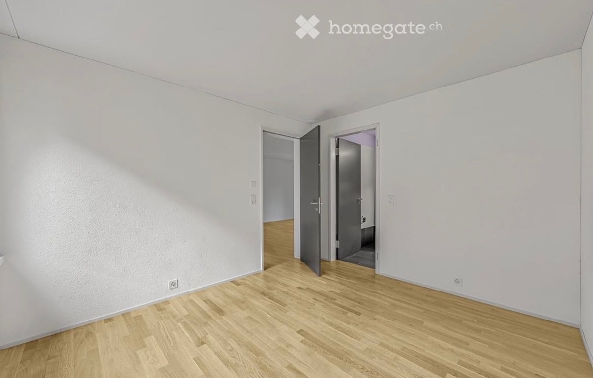 Empty room with light wooden flooring and white walls, featuring two open doors leading to adjacent spaces.