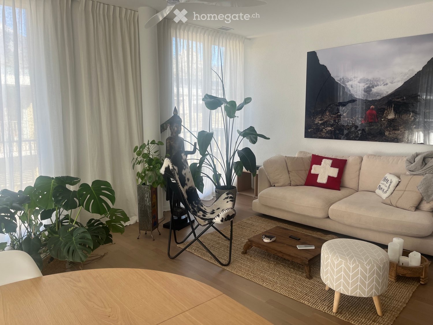Modern living room with a beige sofa, wooden coffee table, patterned stool, and large plants. Features a large window with white curtains and a scenic wall art piece.