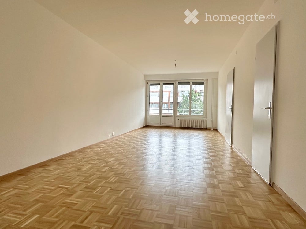 Empty room with wooden herringbone flooring, white walls, large windows, and a door.