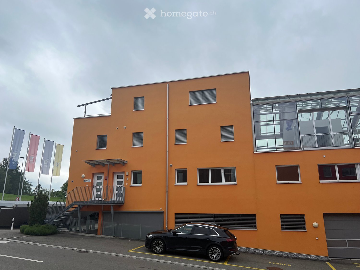 Modern two-story building with orange facade, large windows, and a glass balcony area on the upper floor. Features include a staircase leading to the entrance, a parked car in front, and several flags on poles to the left.