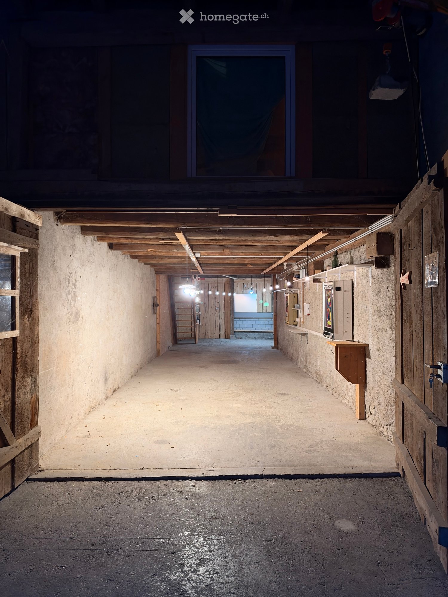 A long, narrow corridor with exposed wooden beams and concrete walls, featuring a series of doors and a small bench on the right side.