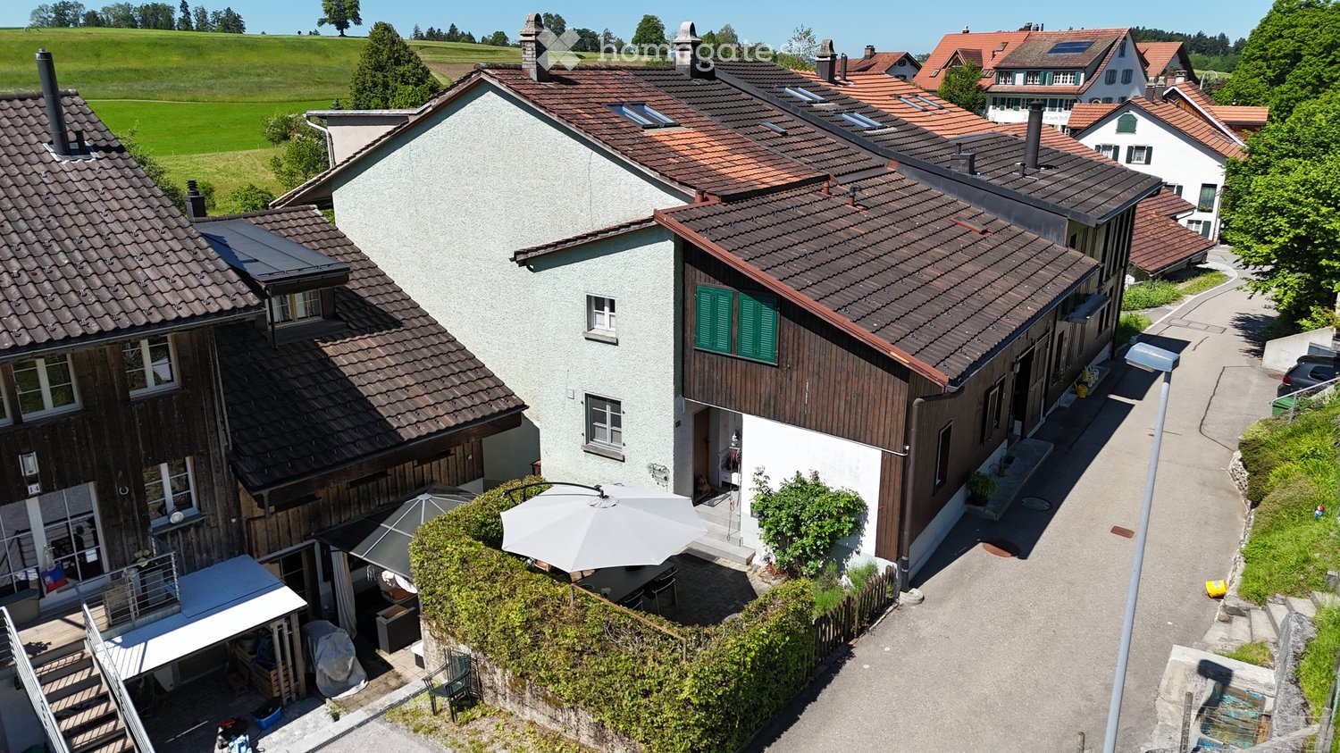 Aerial view of a residential area with multiple houses, featuring red-tiled roofs, green shutters, and a mix of modern and traditional architecture. The houses are surrounded by greenery and a scenic countryside backdrop.