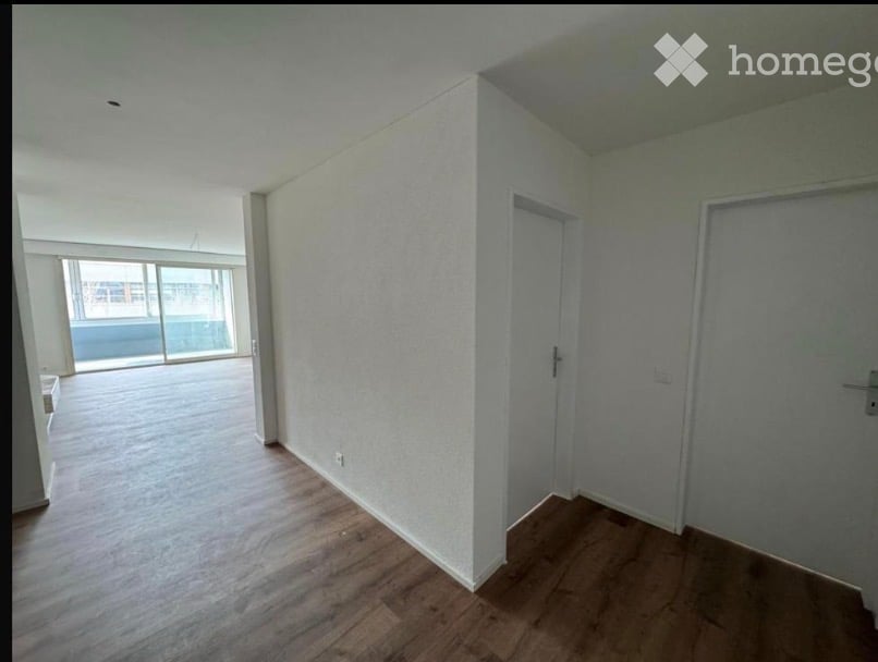 Empty room with wooden flooring, white walls, and large windows offering a view of the outside area.
