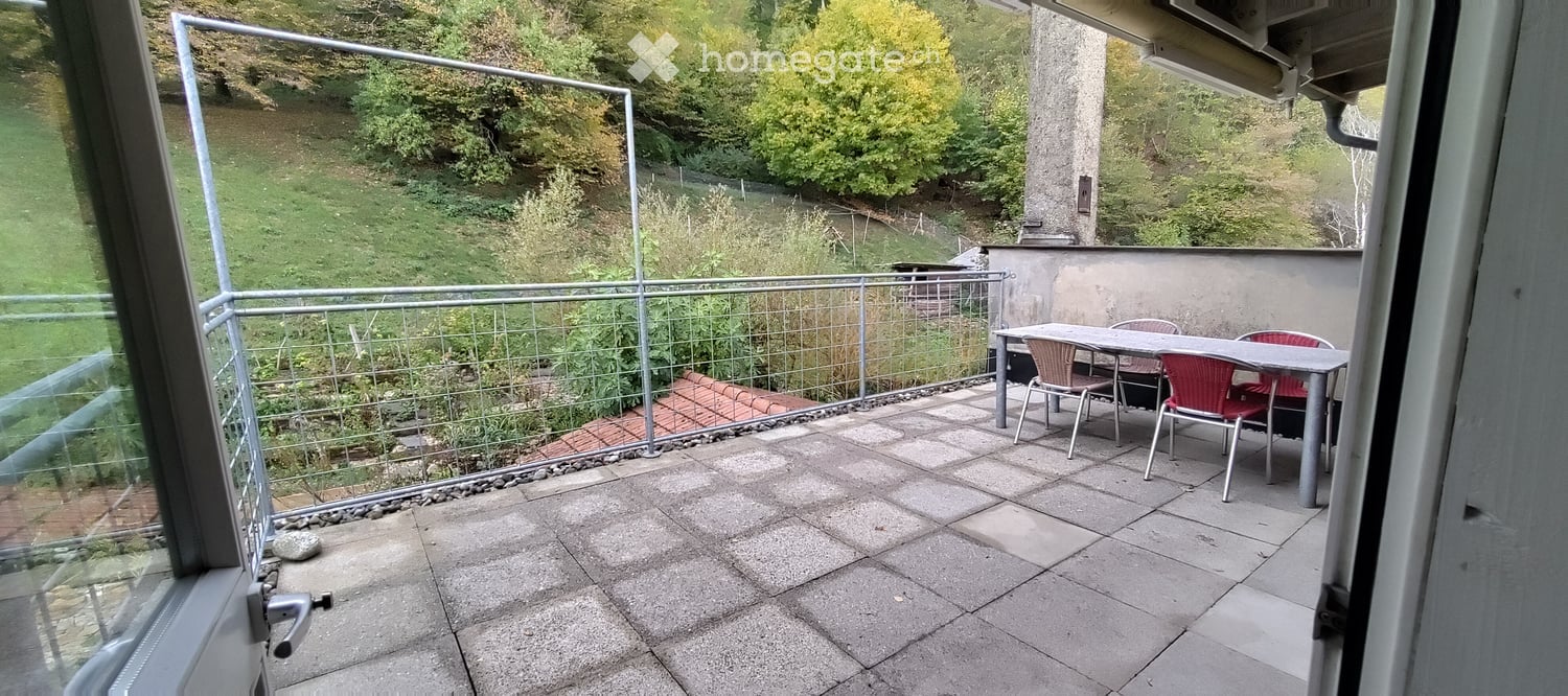Balcony with a table and chairs, overlooking a green, hilly landscape with trees.