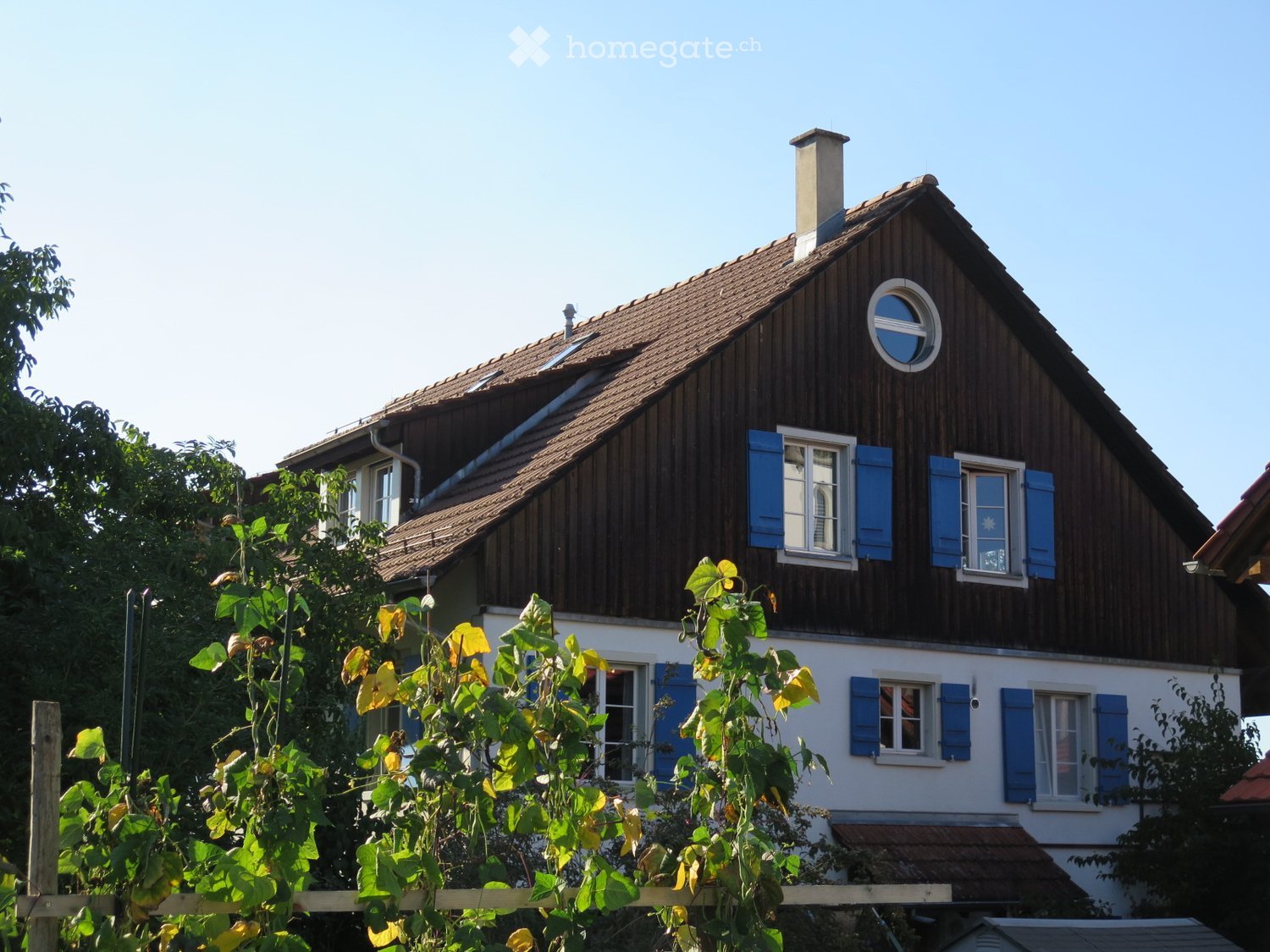 Traditional house with brown wooden exterior, blue shutters, white trim, round window on the roof, solar panels on the roof, brick chimney, surrounded by trees and plants.