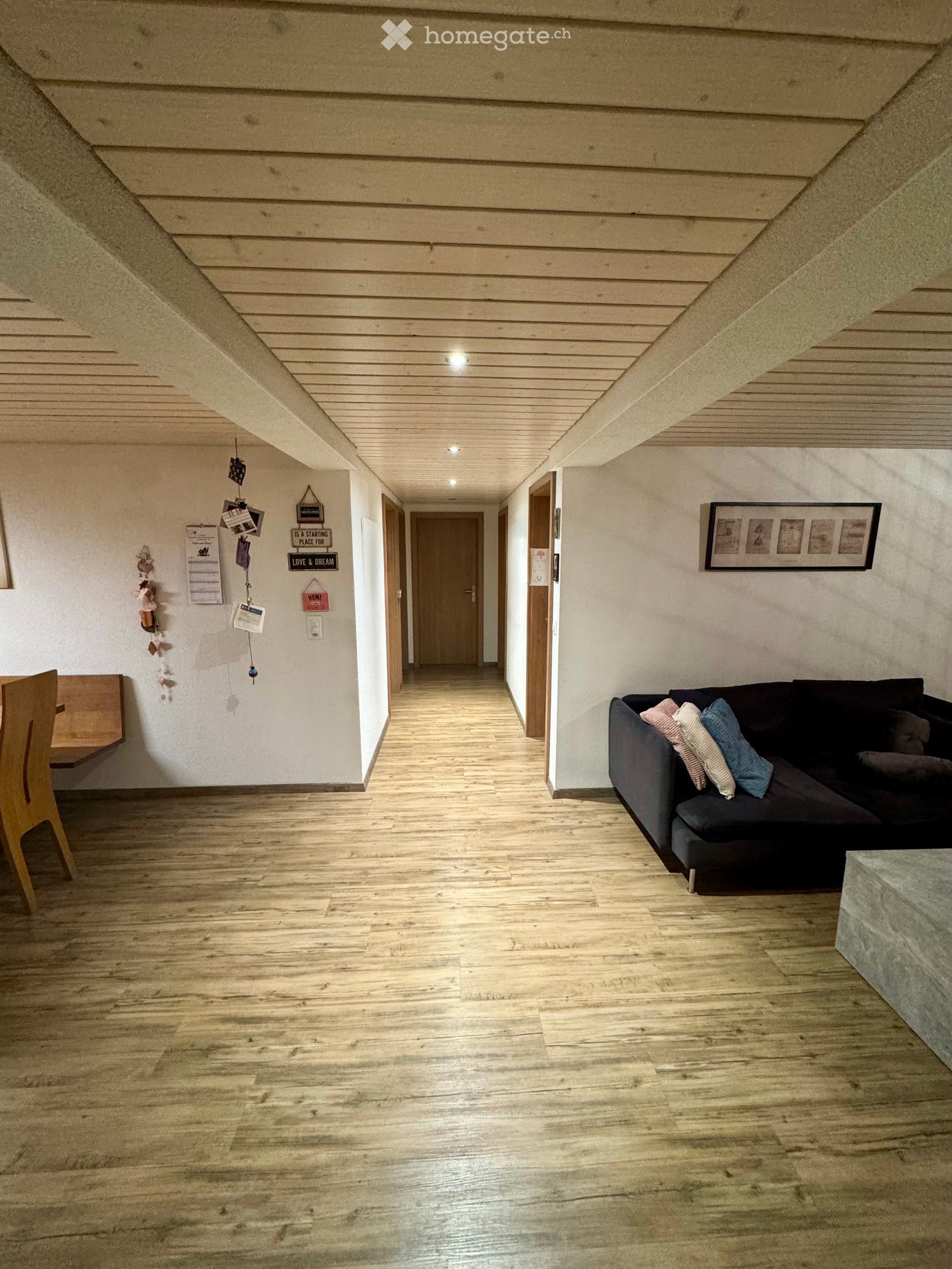 A modern living room with wooden flooring, a dark-colored sofa with pillows, a wooden chair, and a small table. The room features a slanted wooden ceiling and several doors leading to other areas.