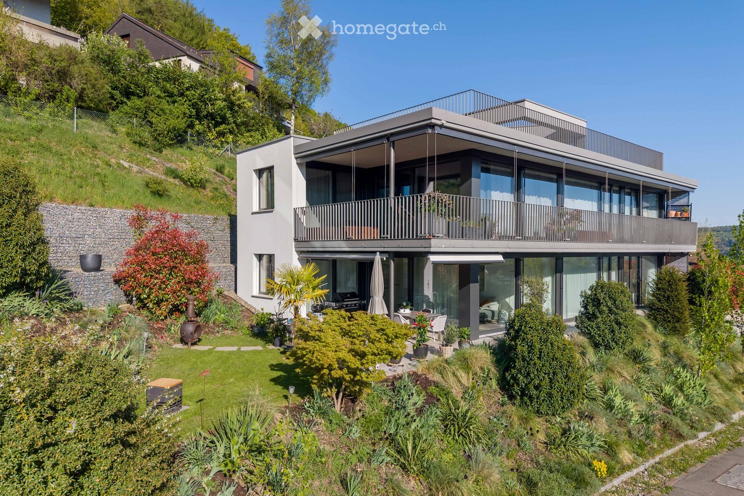 Modern two-story house with large windows and a balcony, surrounded by a well-maintained garden with various plants and trees.