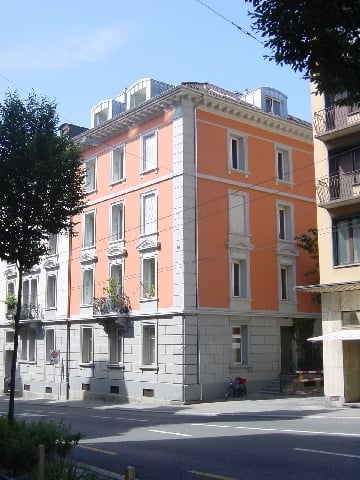 Four-story apartment building with orange and white facade, multiple windows, balconies, and greenery on the ground floor.