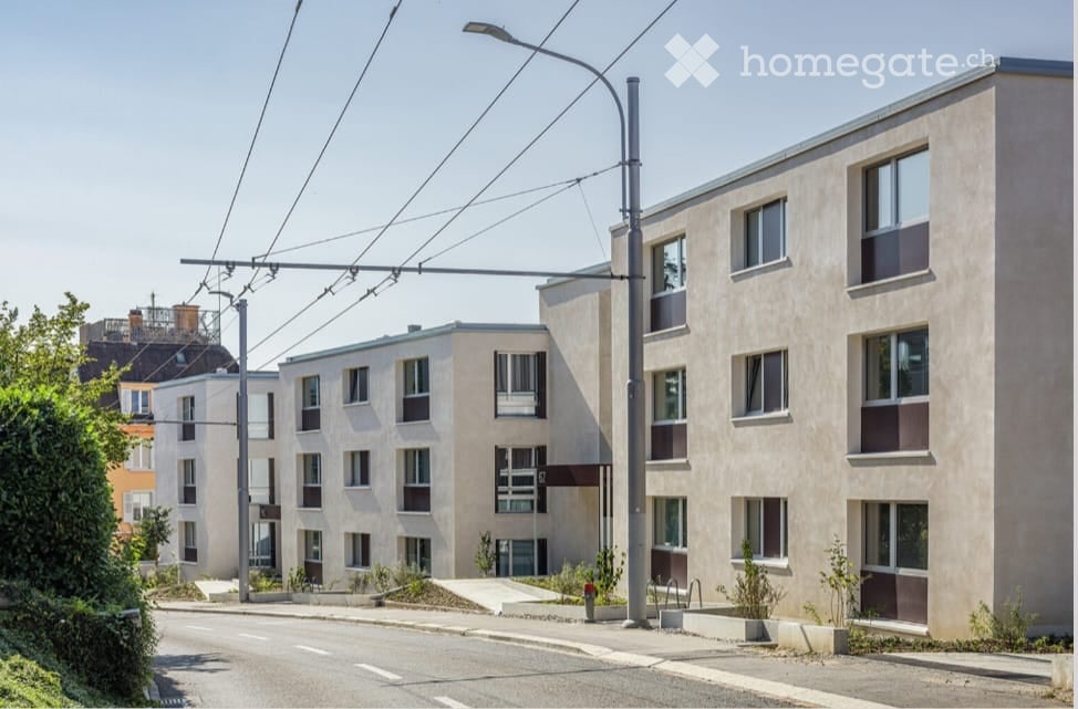 Modern multi-story residential building with beige facade, multiple windows, and small landscaped areas in front.