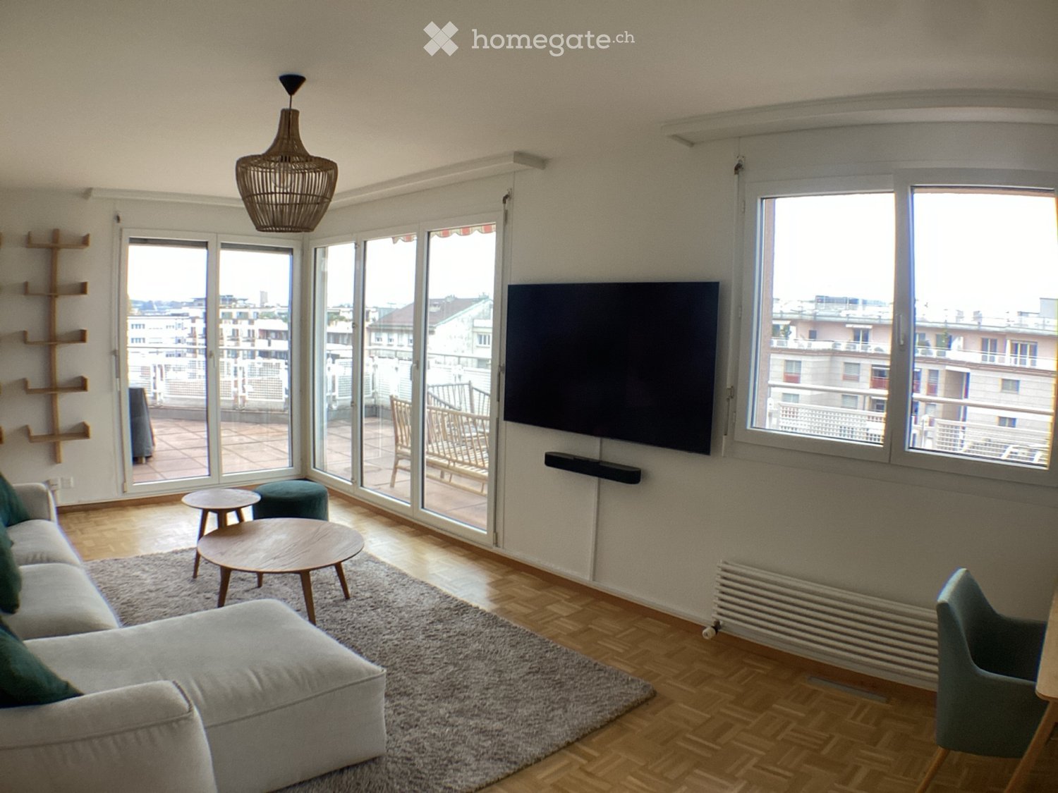 Modern living room with large windows and a balcony, featuring a grey sofa, coffee table, wooden chairs, and a television.