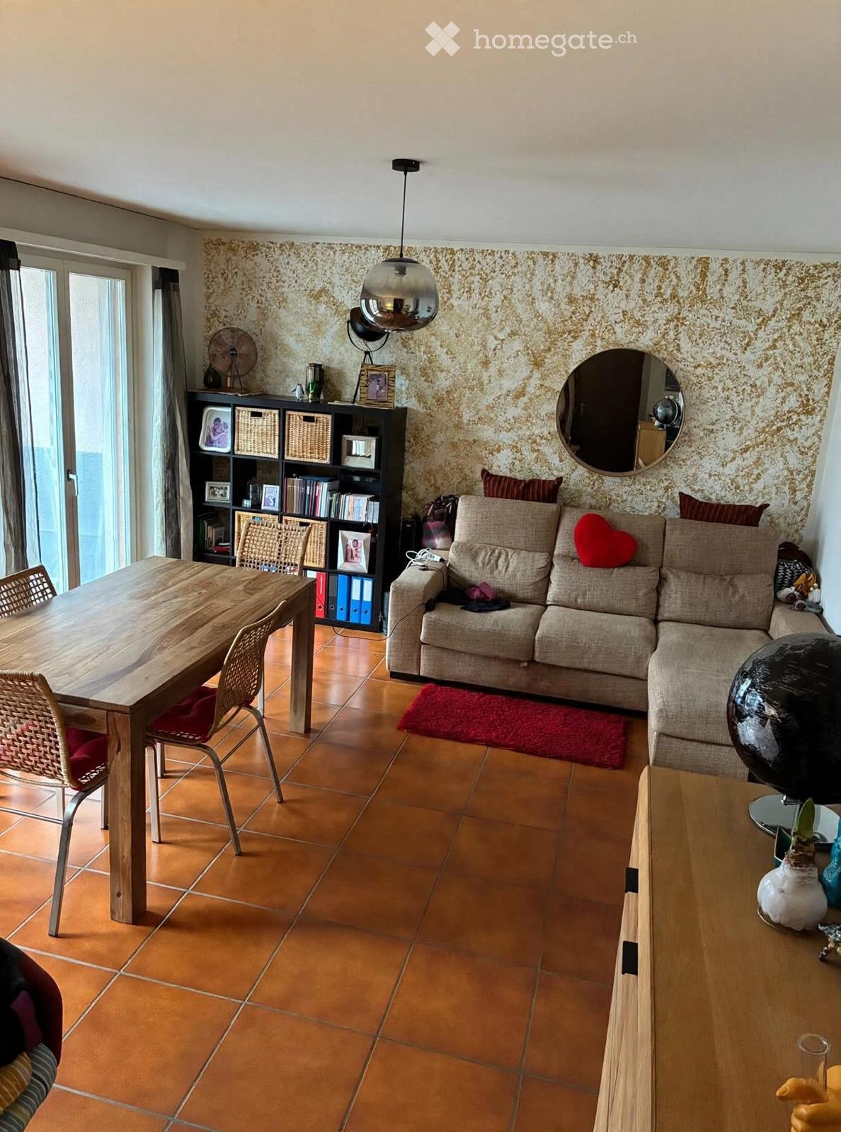 Living room with a beige sofa, wooden coffee table, dining table with chairs, bookshelf, mirror, fan, globe, and decorative items.