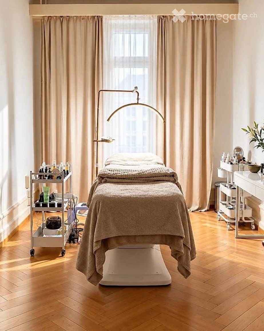 A well-lit, modern massage room with a massage table, a cart with various items, a large window with curtains, a floor lamp, and a small plant on a side table.