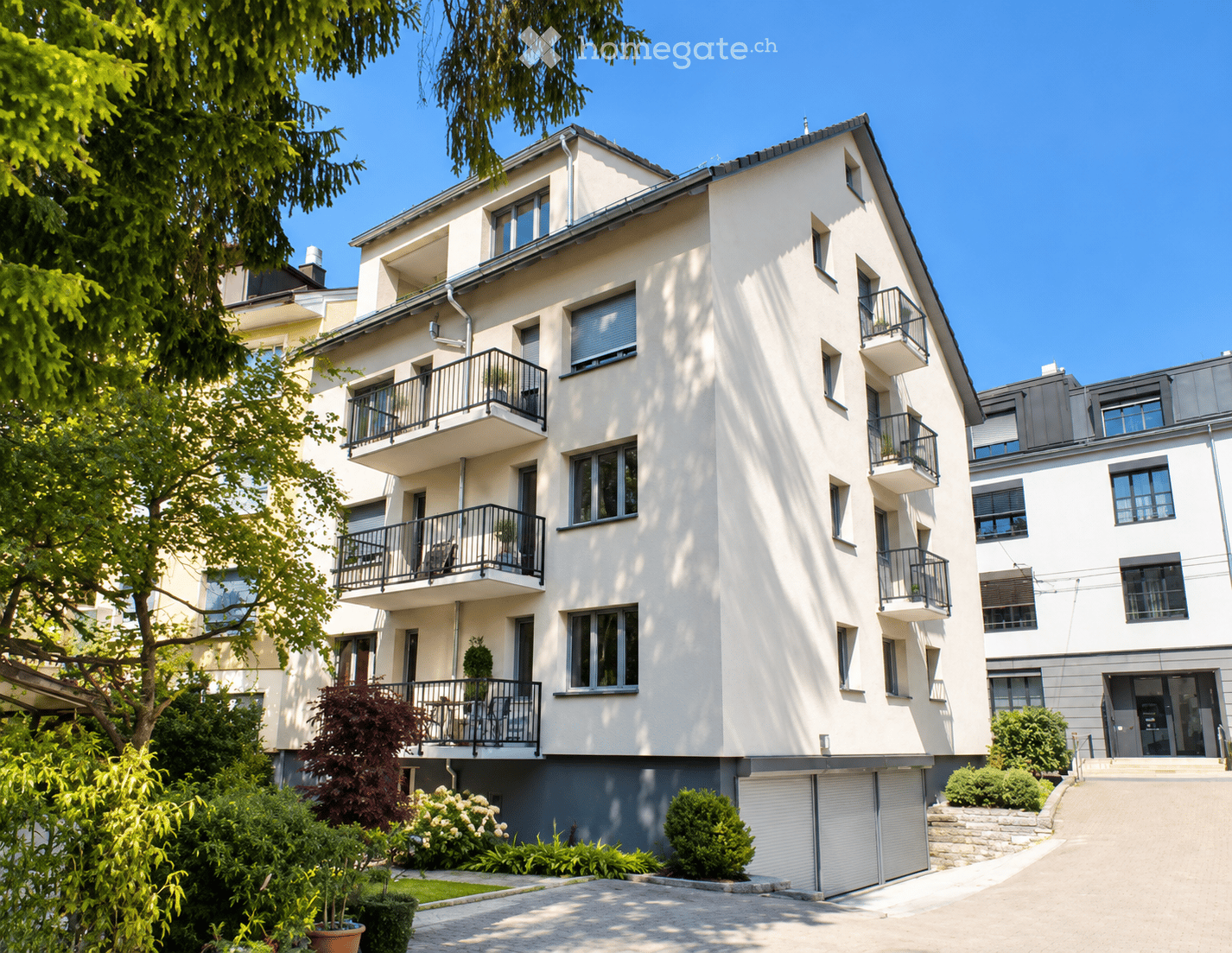 Modern multi-story residential building with white facade, multiple balconies, and a garage entrance.