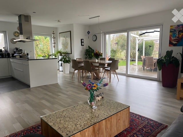 Open-plan living and dining area with modern furniture, large sliding glass doors leading to a garden, and a kitchen area with cabinets and a range hood.