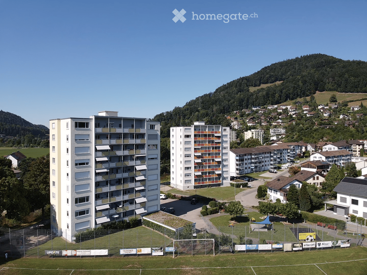 Aerial view of a residential area with multiple apartment buildings, surrounded by green spaces, trees, and a small sports field.