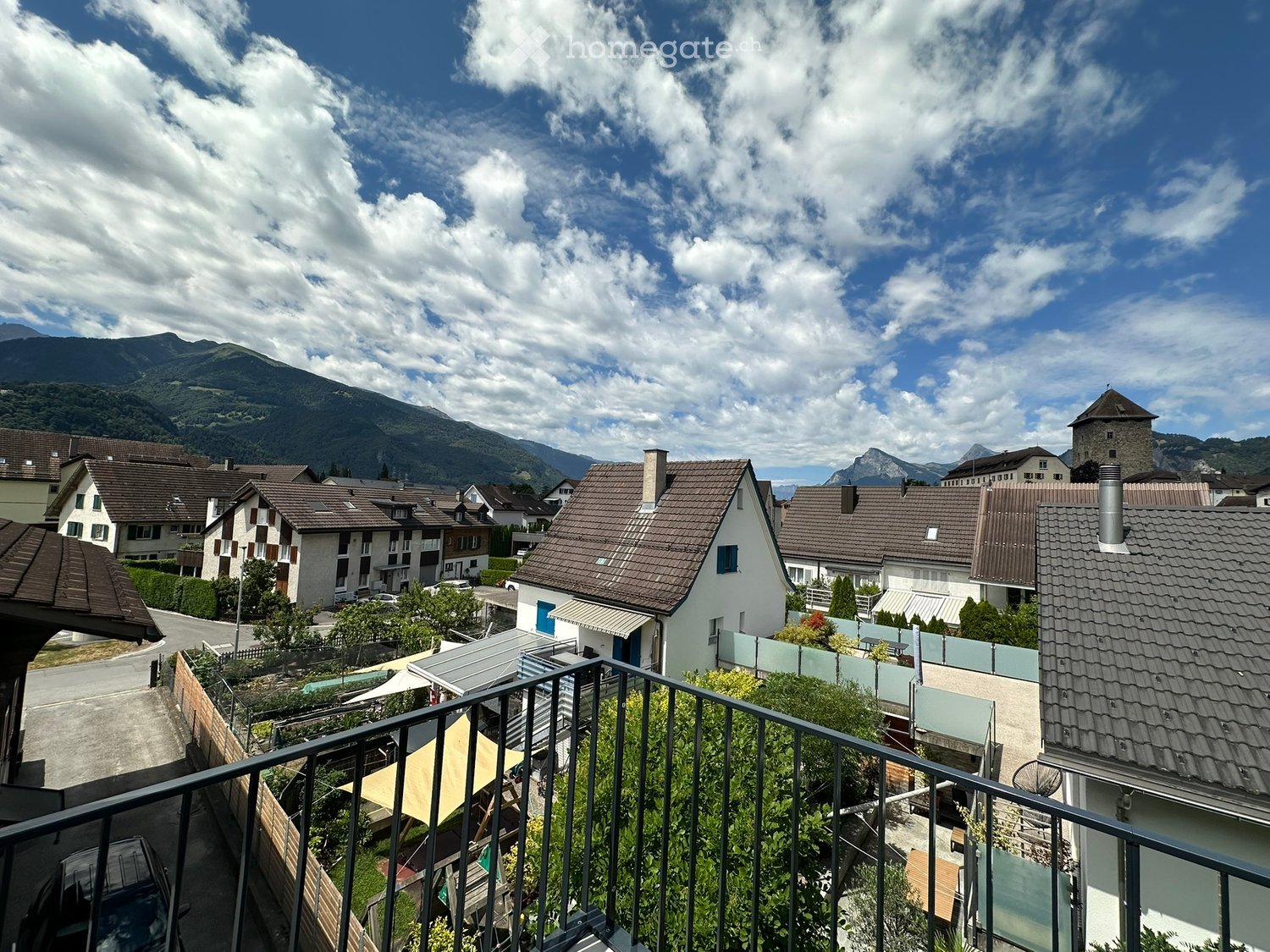 Balcony overlooking a residential area with houses, mountains, and a partly cloudy sky.