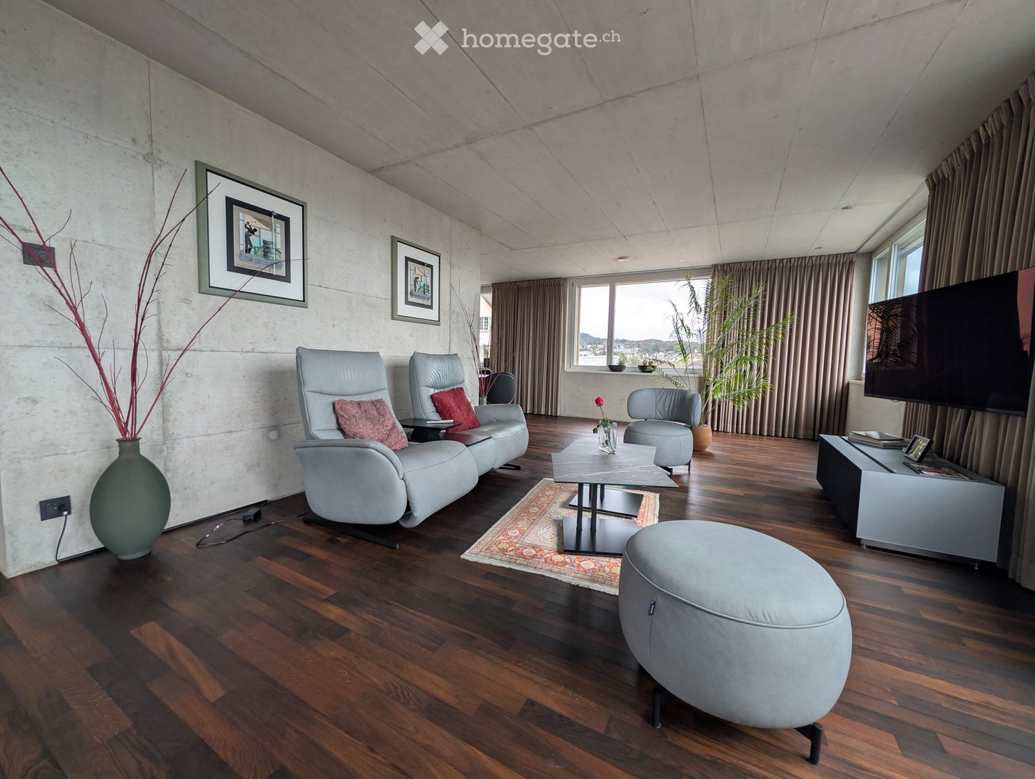 Modern living room with wooden flooring, grey leather sofa, coffee table, ottoman, chairs, vase, television, and picture frames.