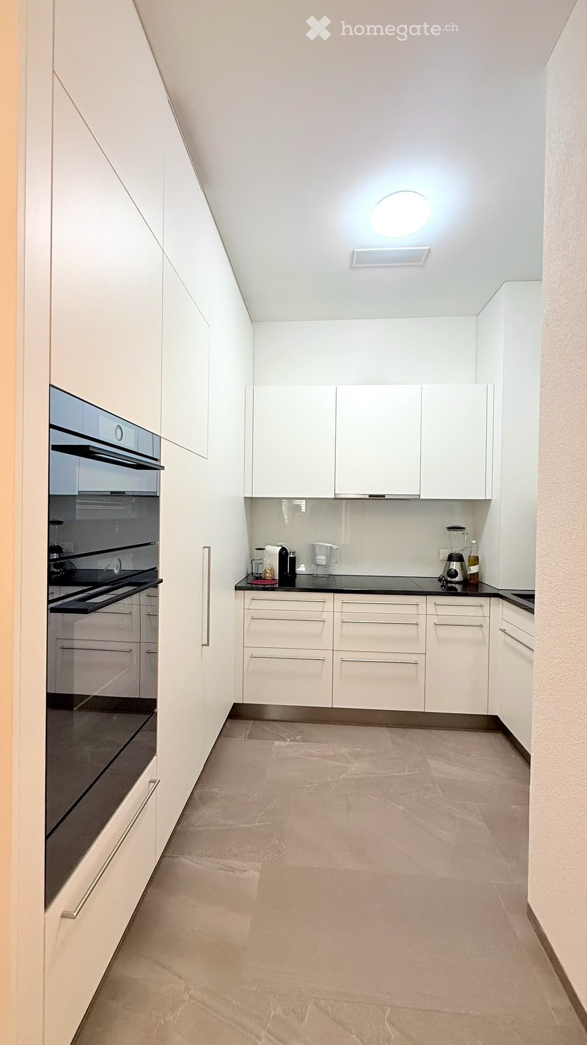 Modern kitchen with white cabinets, black countertops, and a built-in oven. The space features a sleek design with a light-colored tiled floor and a round ceiling light.