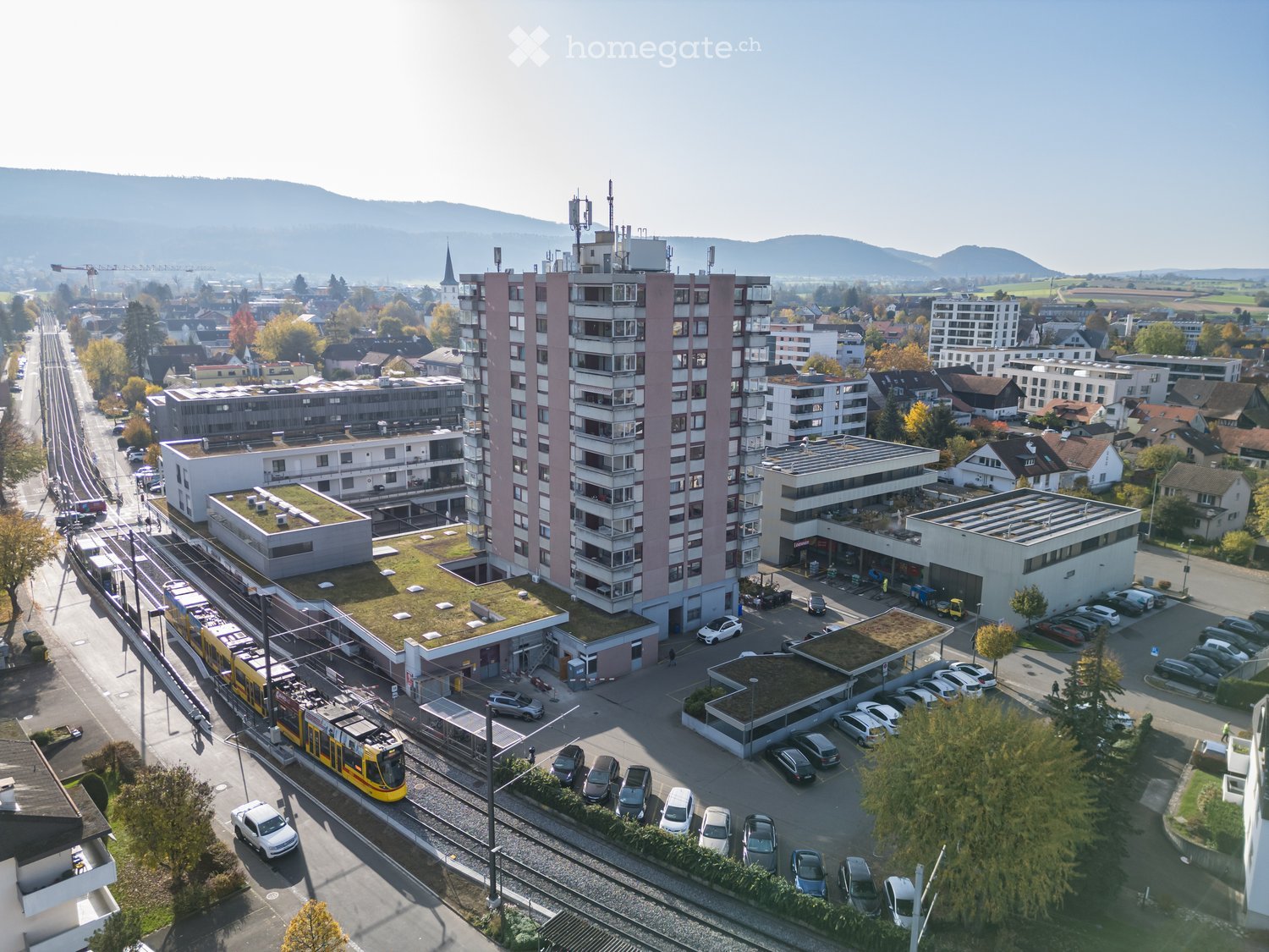 Aerial view of a multi-story building with green roofs, surrounded by other buildings, a train track, and a parking lot with many cars.