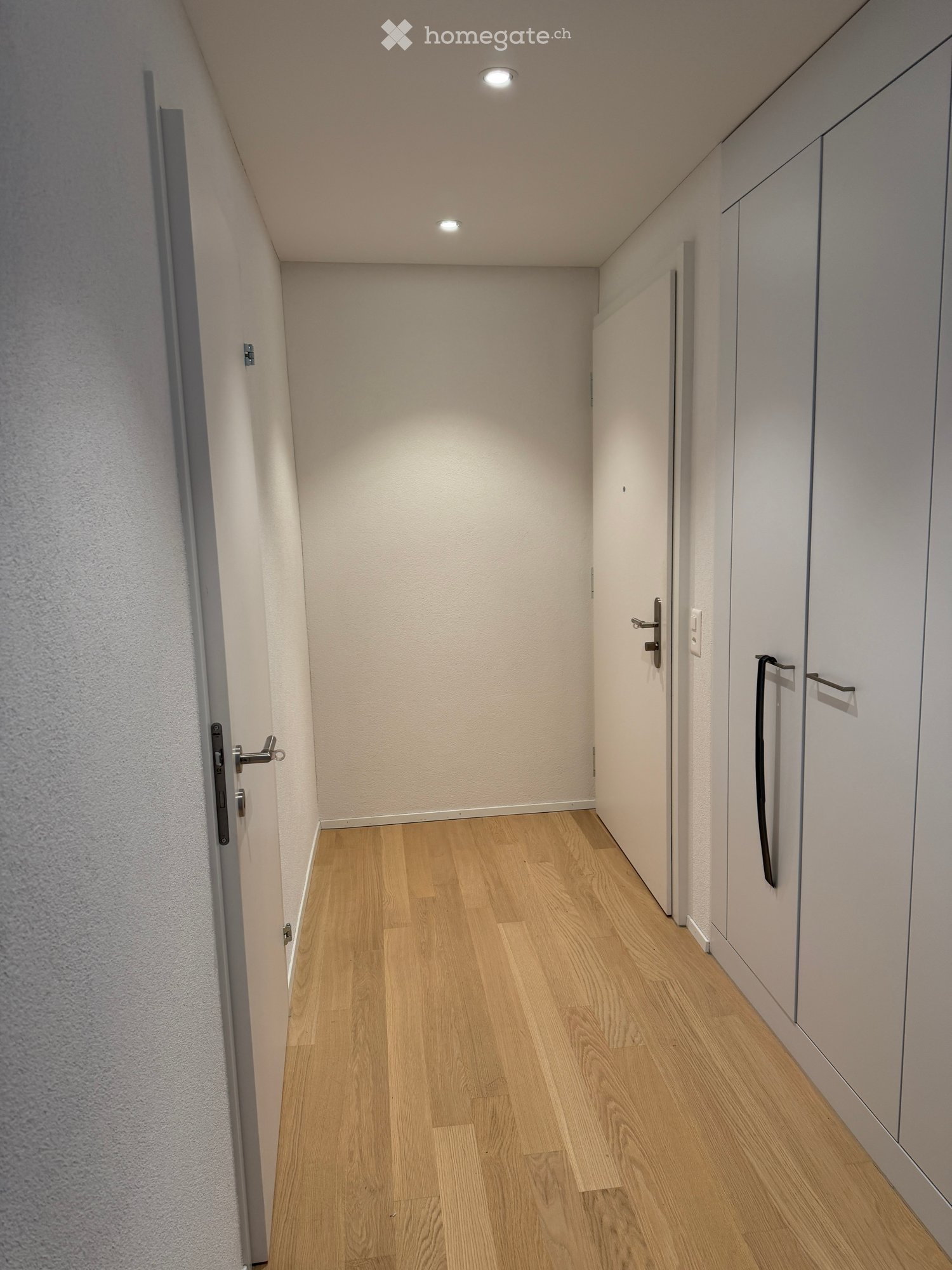 Narrow hallway with light wooden flooring, white walls, and built-in cabinets on one side.