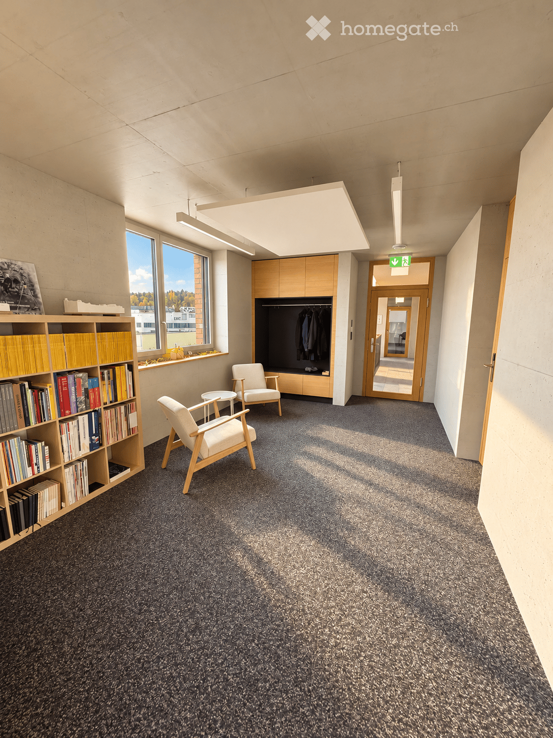 Modern, well-lit room with grey carpet, white chairs, small round table, wooden bookshelf filled with books, and a cabinet with coats hanging inside.