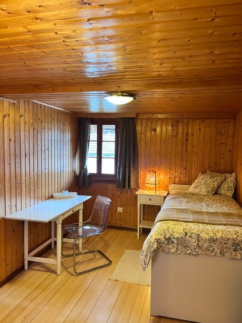 Cozy wooden bedroom with a double bed, nightstand with lamp, small desk, and chair. Wooden walls and ceiling, warm lighting, and a window with dark curtains.