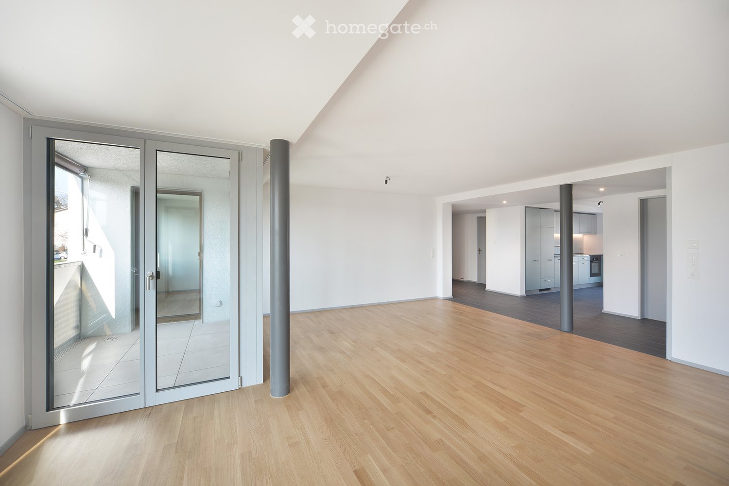 Bright, empty room with wooden flooring, white walls, and large glass sliding doors leading to a balcony. The space is clean and minimalistic, with no furniture or personal items visible.