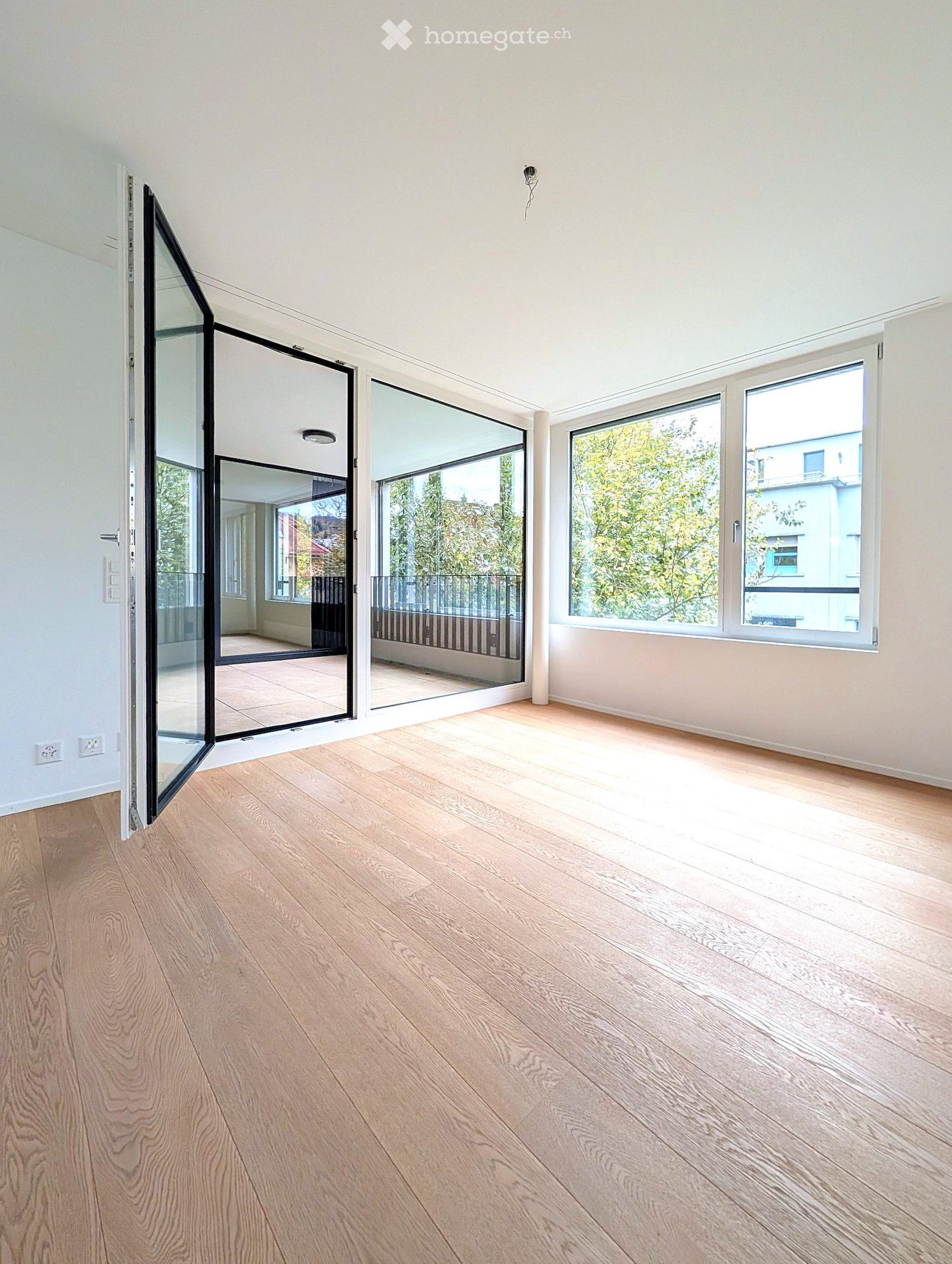Empty room with light wooden flooring, white walls, large windows, and a set of glass folding doors leading to a balcony.