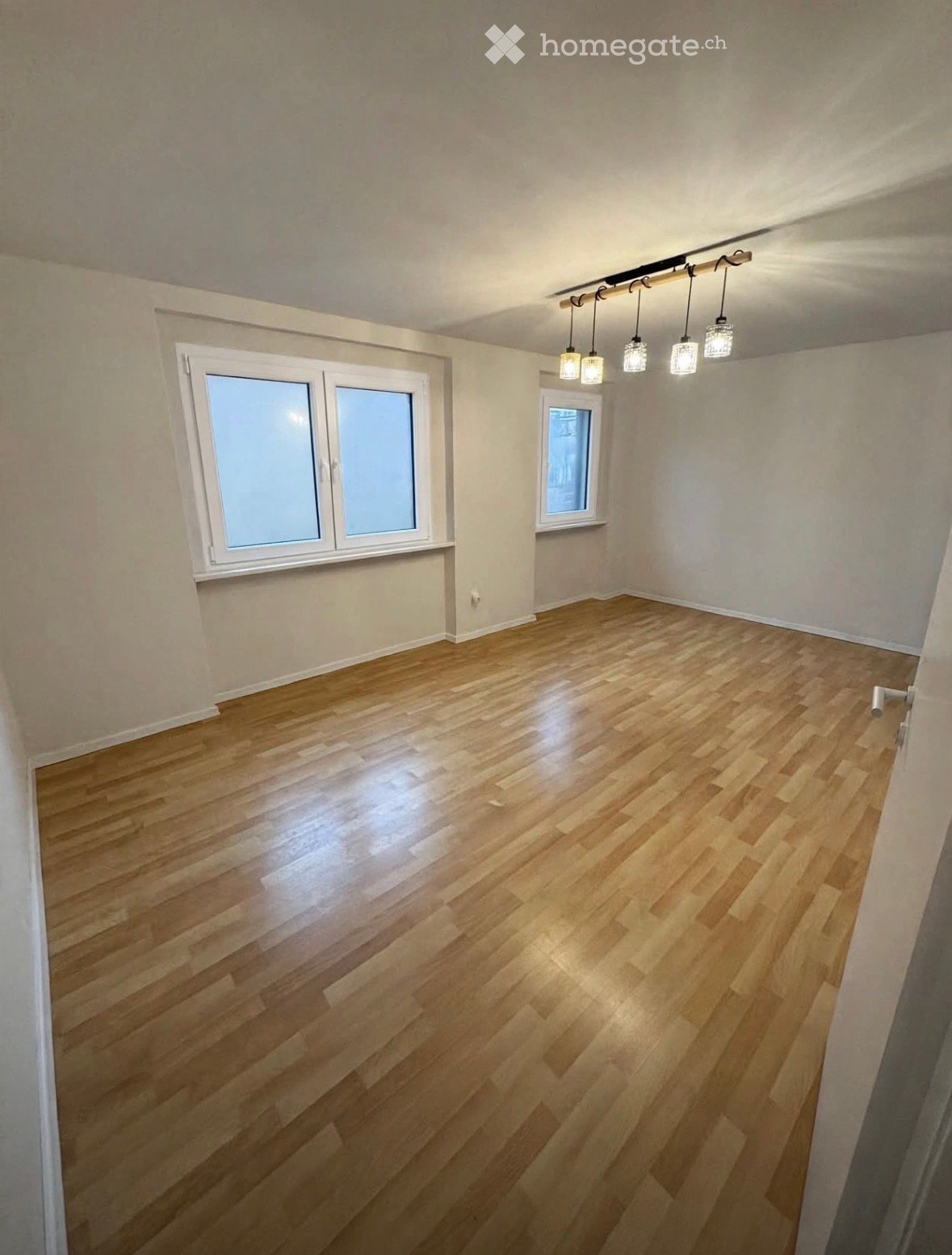 Empty room with wooden flooring, white walls, and large windows. The room features modern lighting fixtures suspended from the ceiling.
