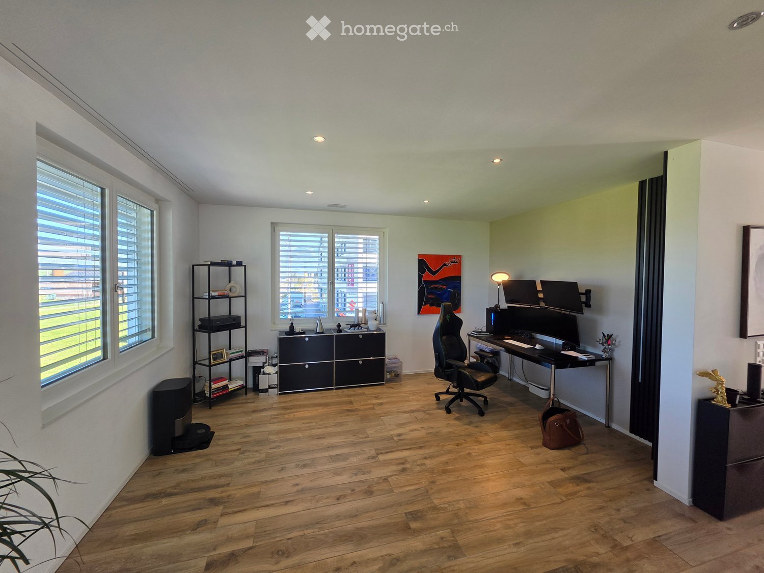 Modern office space with wooden flooring, large windows with blinds, a black office chair, desk with dual monitors, a floor lamp, a black shelving unit, and a cabinet.