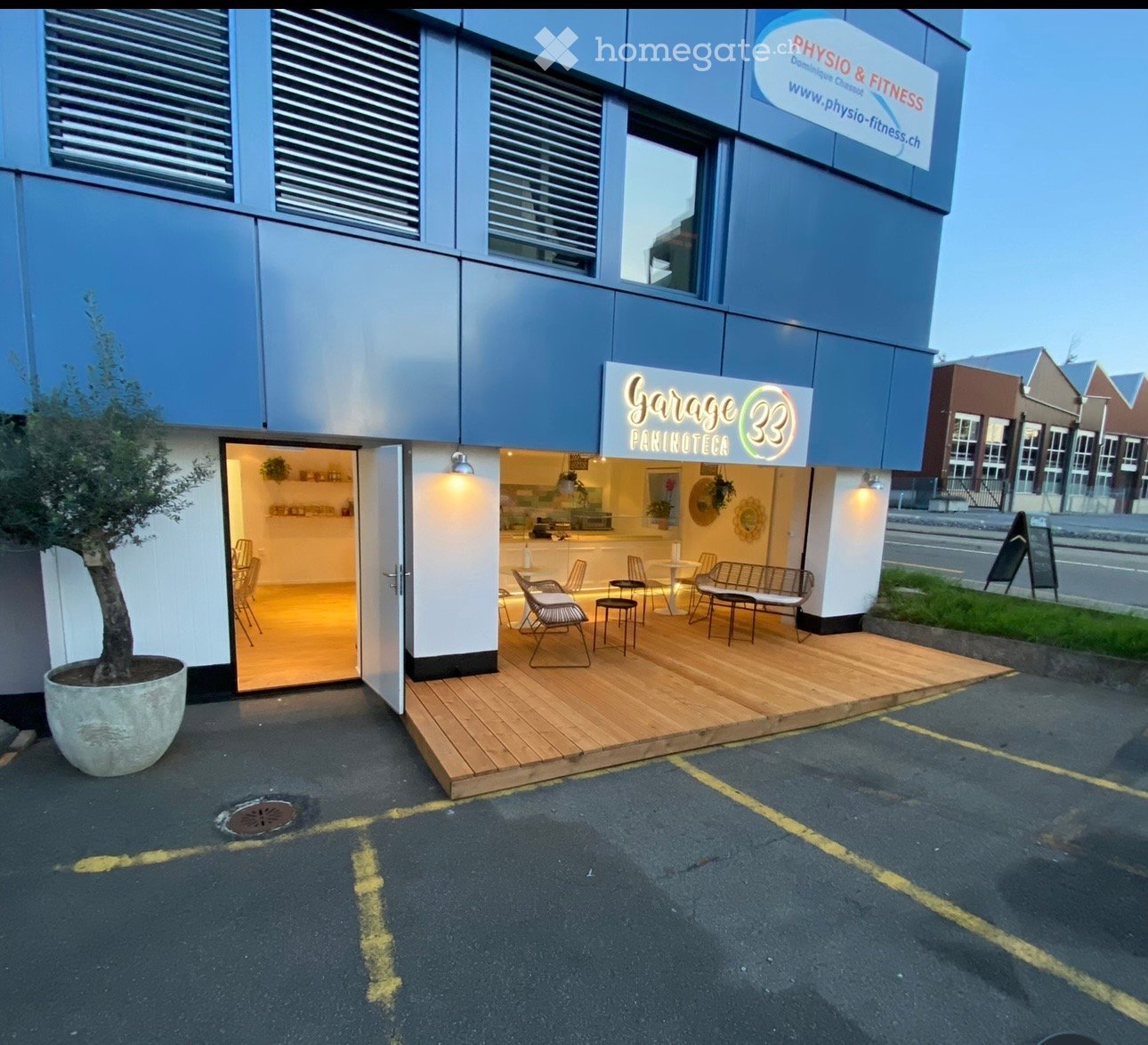 Modern building exterior with a wooden deck area featuring chairs and tables, a potted tree, and signage for "Garage 33 Pakinoteca" and "Physio & Fitness".