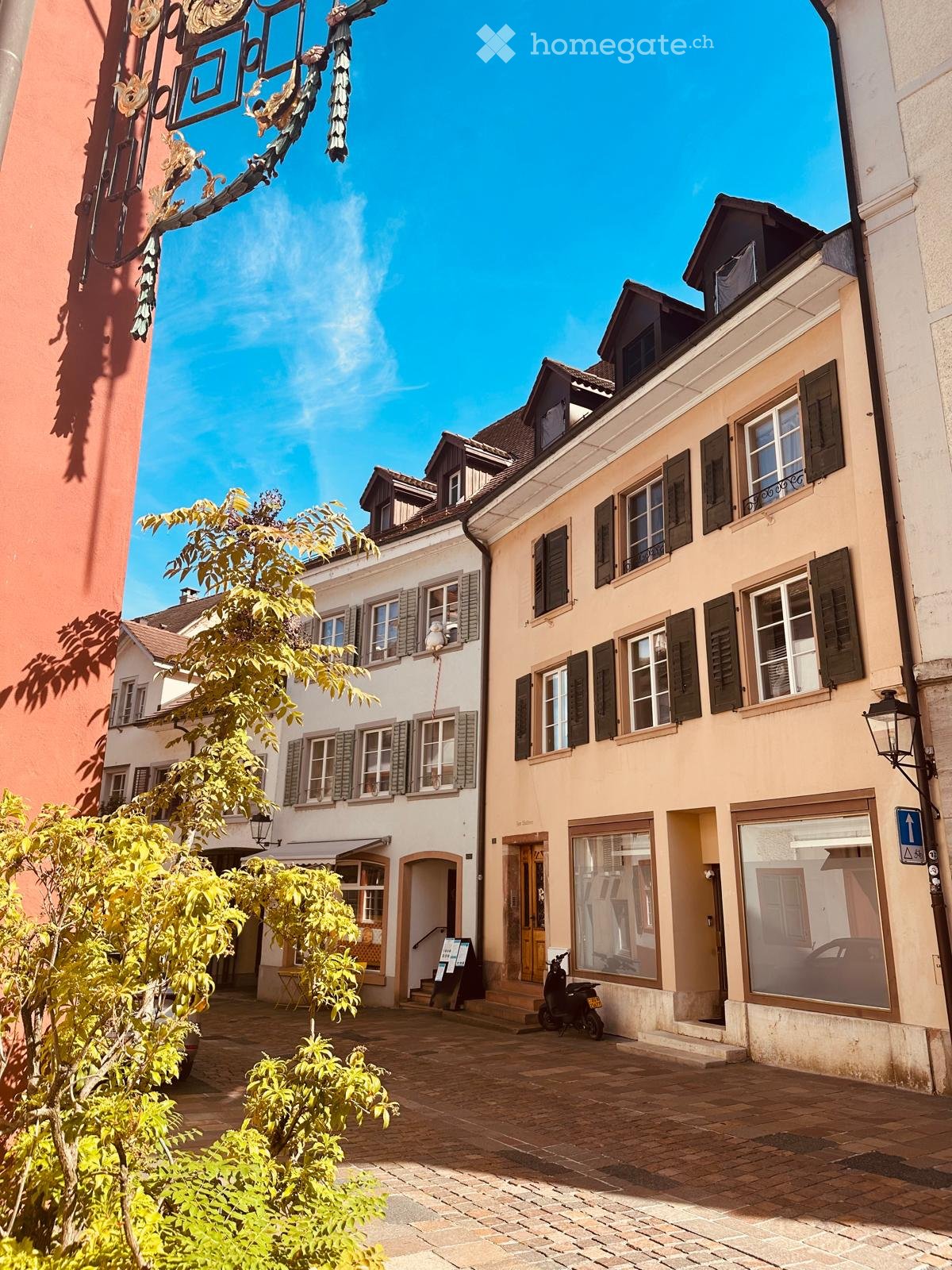 A charming European street scene with classic buildings, cobblestone pavement, and greenery. The buildings feature traditional architecture with shutters and pitched roofs.