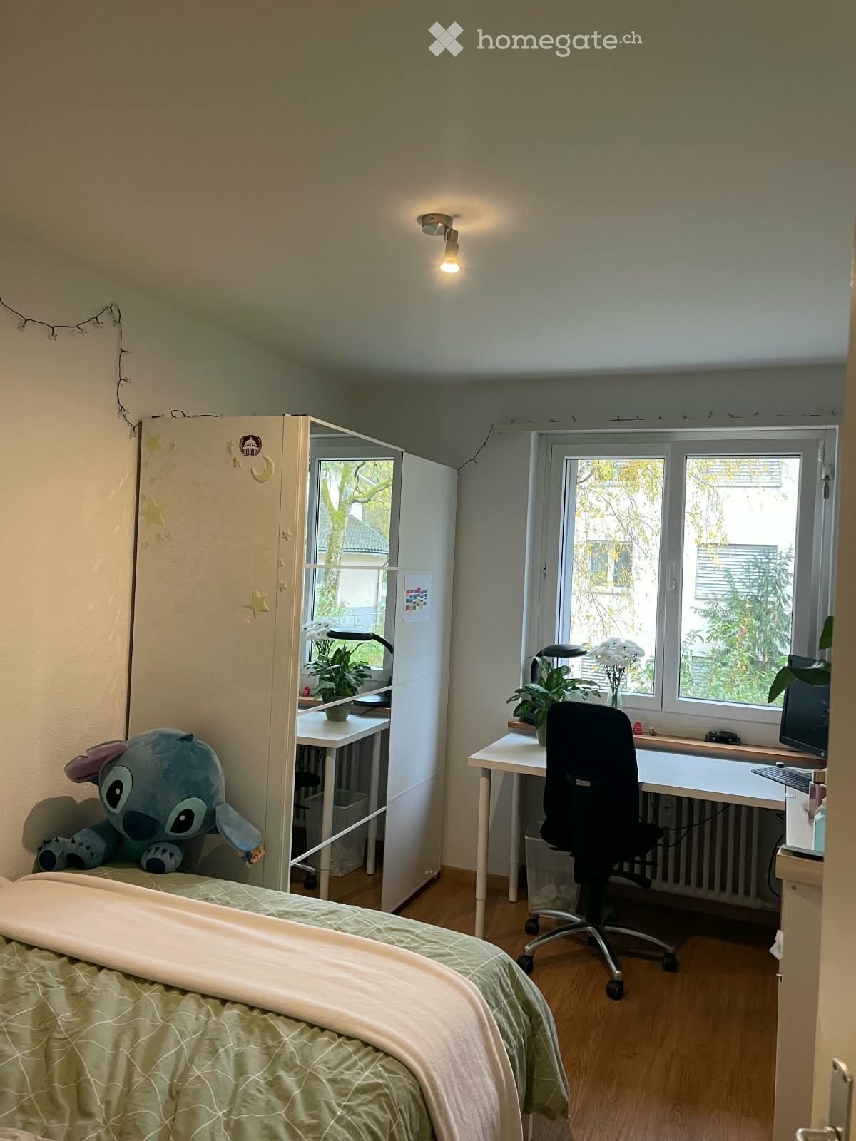 A small, well-lit bedroom with a bed, desk, and chair. The room features a large window with a view of greenery, a wardrobe, and a small plant on the desk.
