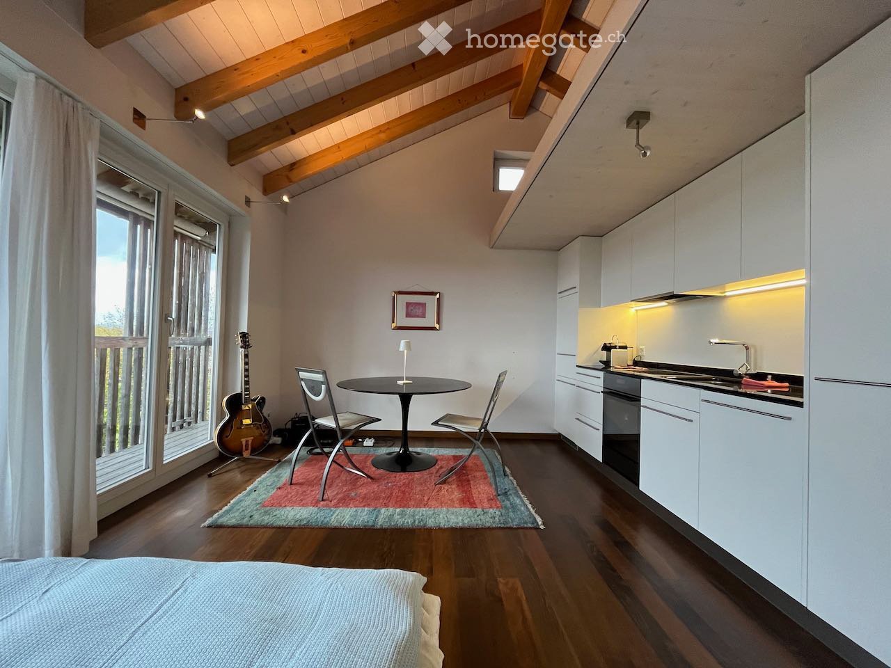 A modern, open-concept room with a bed, a small dining area, and a compact kitchen. Features include wooden beams on the ceiling, large windows with white curtains, and built-in white kitchen cabinets.