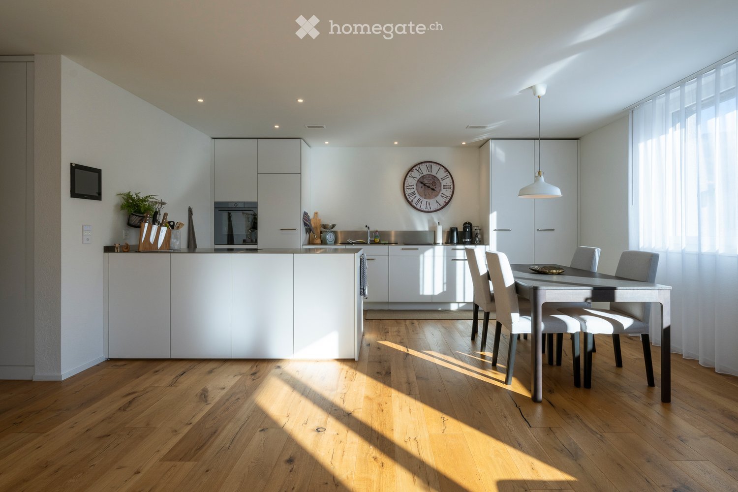 Modern kitchen with white cabinets, a central island, and a dining area with a table and chairs. Features include a clock on the wall, a screen, and a plant on the counter.