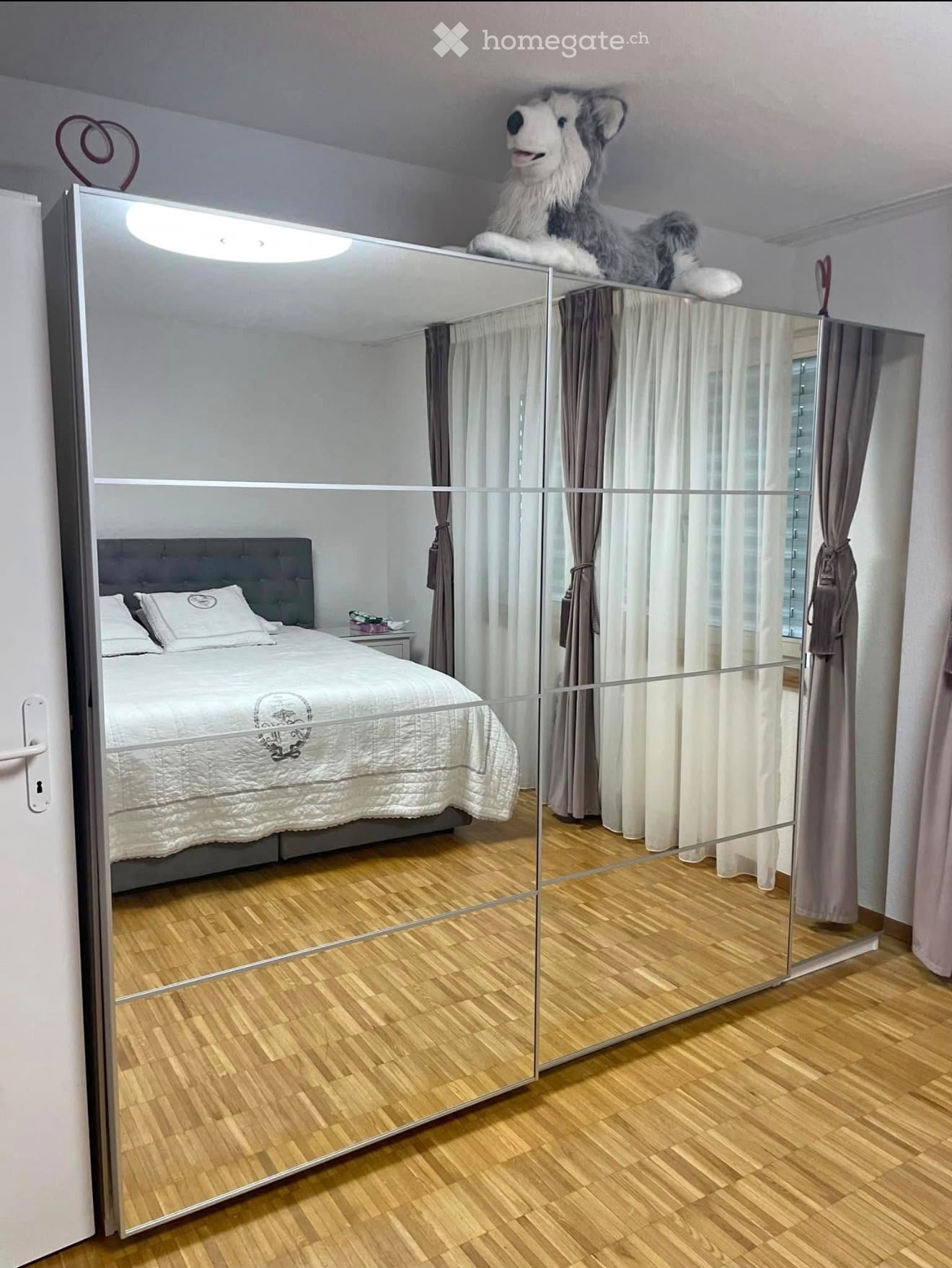 Modern bedroom with a large bed, nightstand, pillows, and a large mirror reflecting the room. The floor is wooden, and there is a plush toy on top of the mirror.