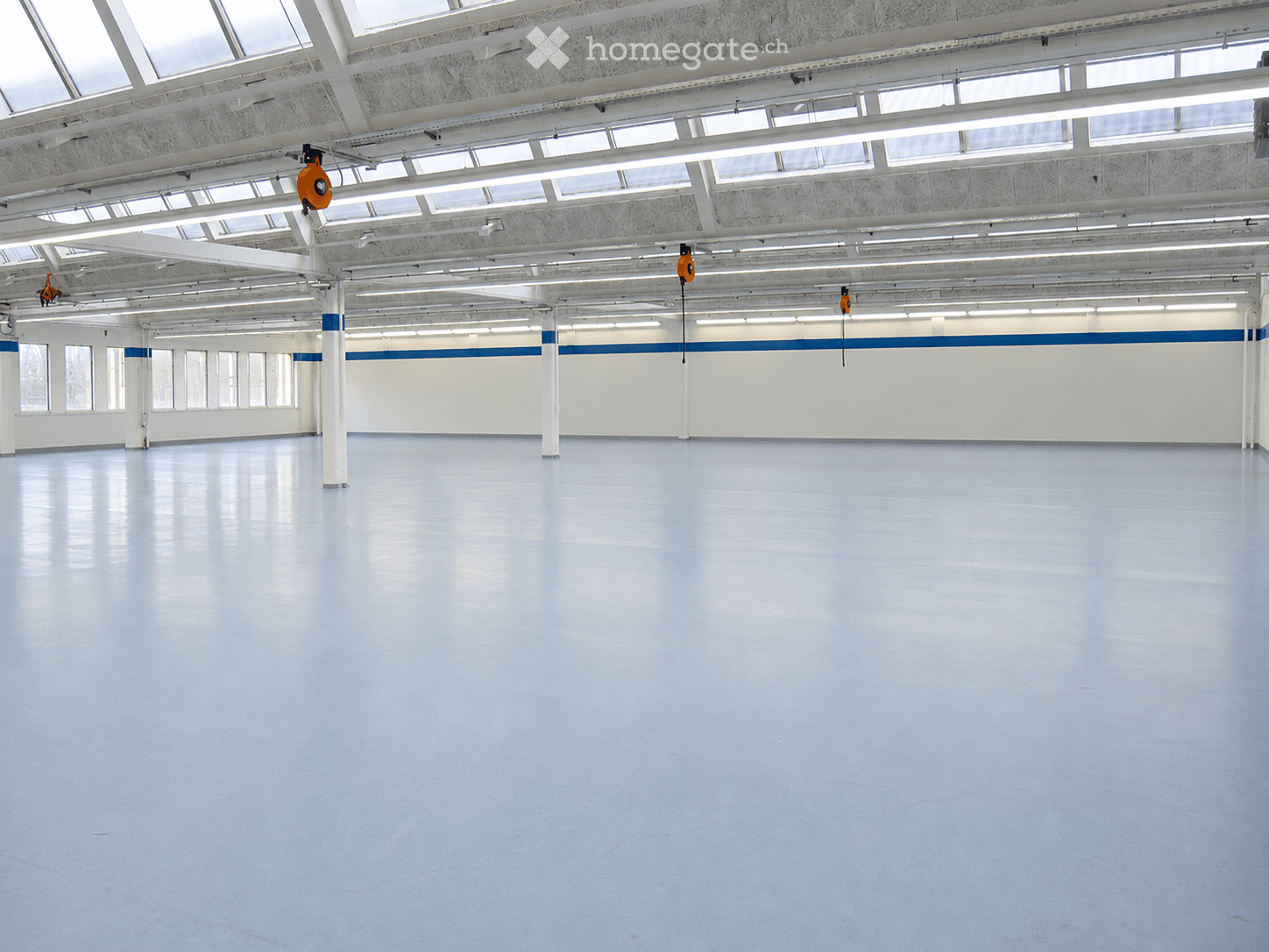 Large, empty industrial-style room with white walls, polished blue floor, and exposed white structural beams and lighting. No furniture or equipment present.